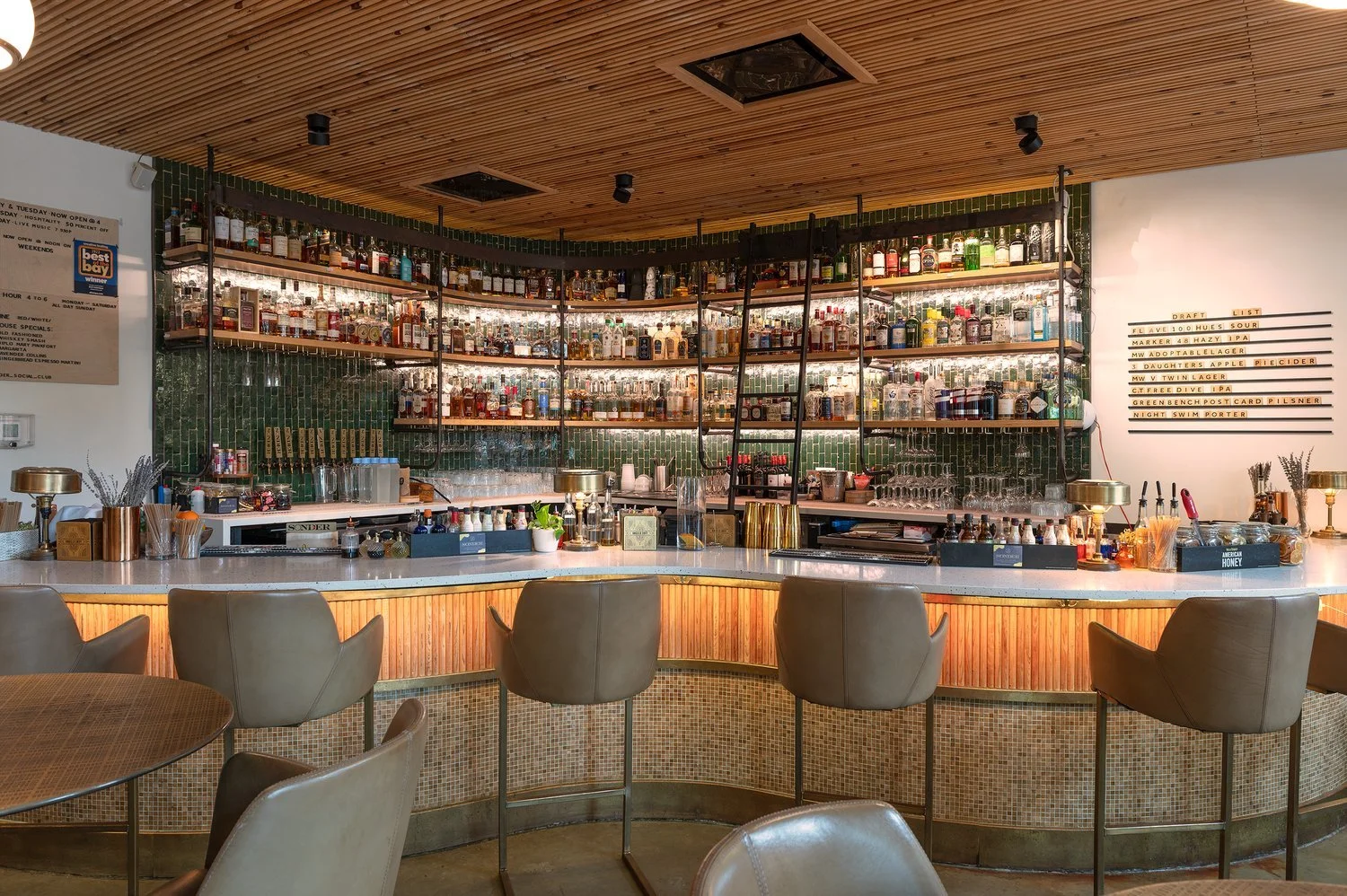 Bar with shelves of liquor bottles, bar stools, and a chalkboard menu listing drinks.