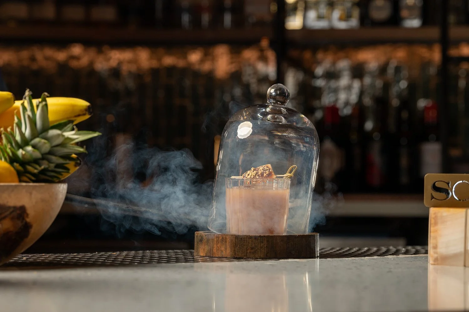 A cocktail topped with a cookie slice under a glass cloche on a wooden coaster, with smoke surrounding it, next to a bowl of bananas.