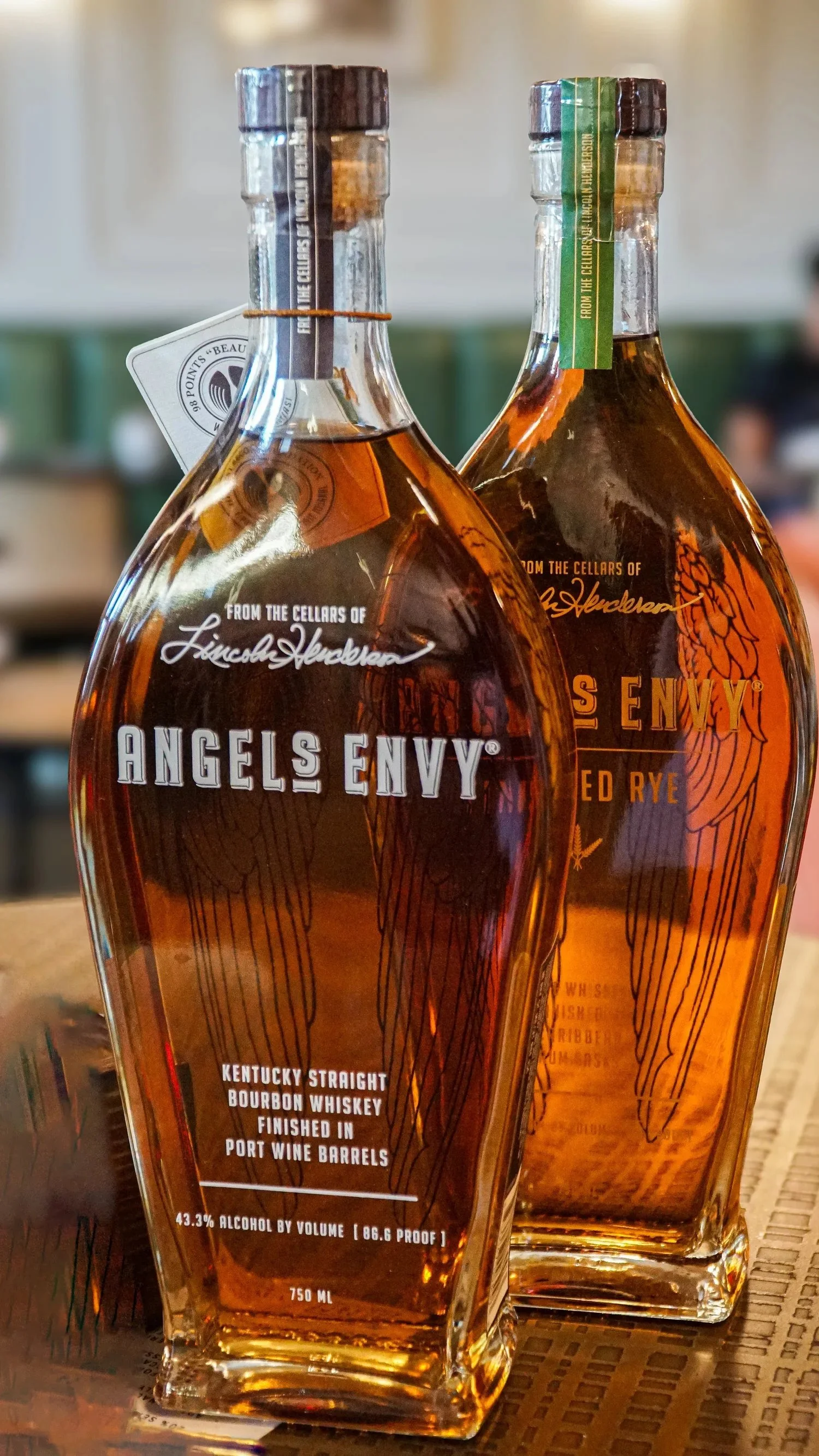 Two bottles of Angels Envy Kentucky Straight Bourbon Whiskey on a wooden surface with blurred background.