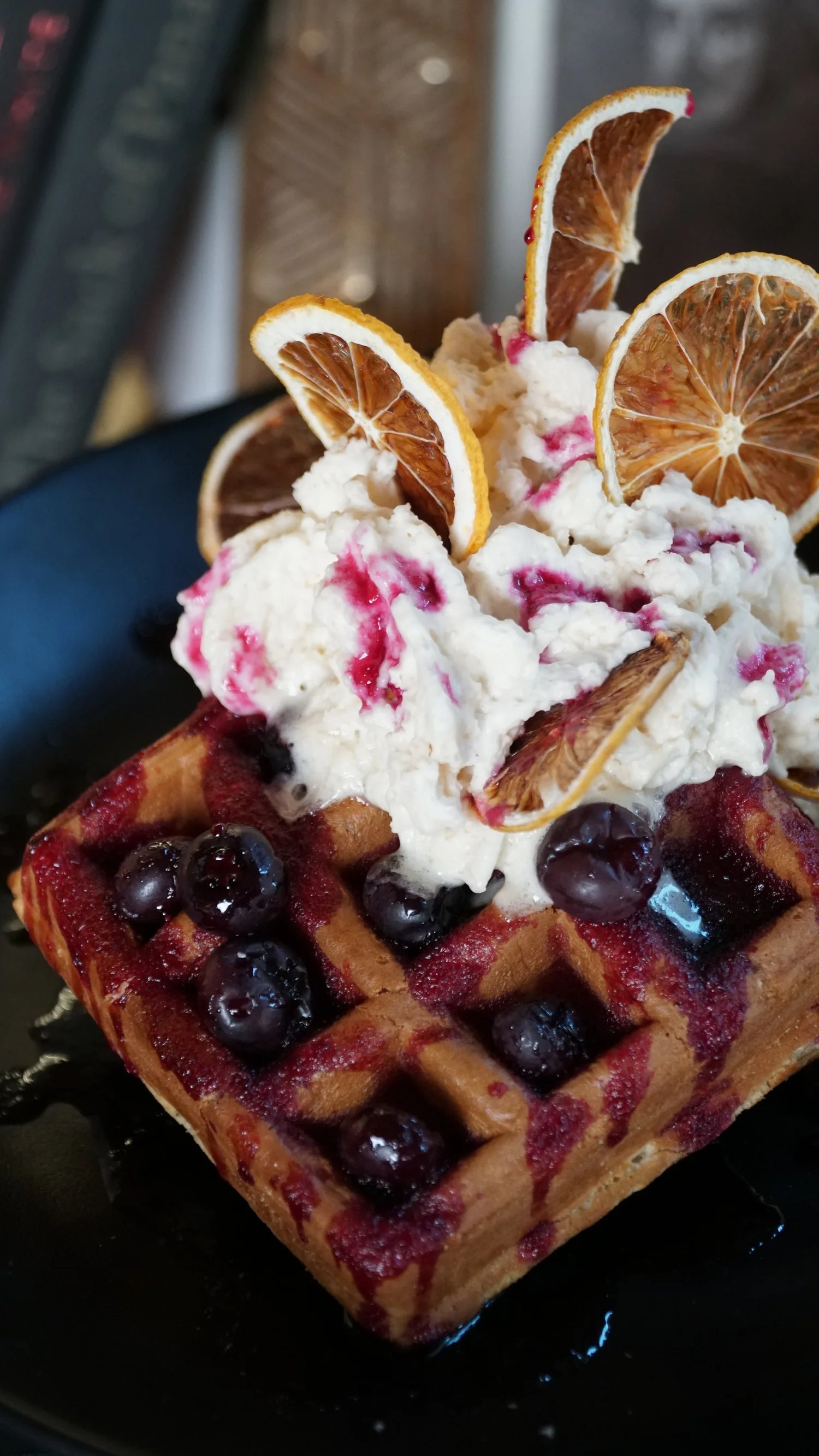 A waffle topped with blueberries, whipped cream, dried citrus slices, and berry syrup.