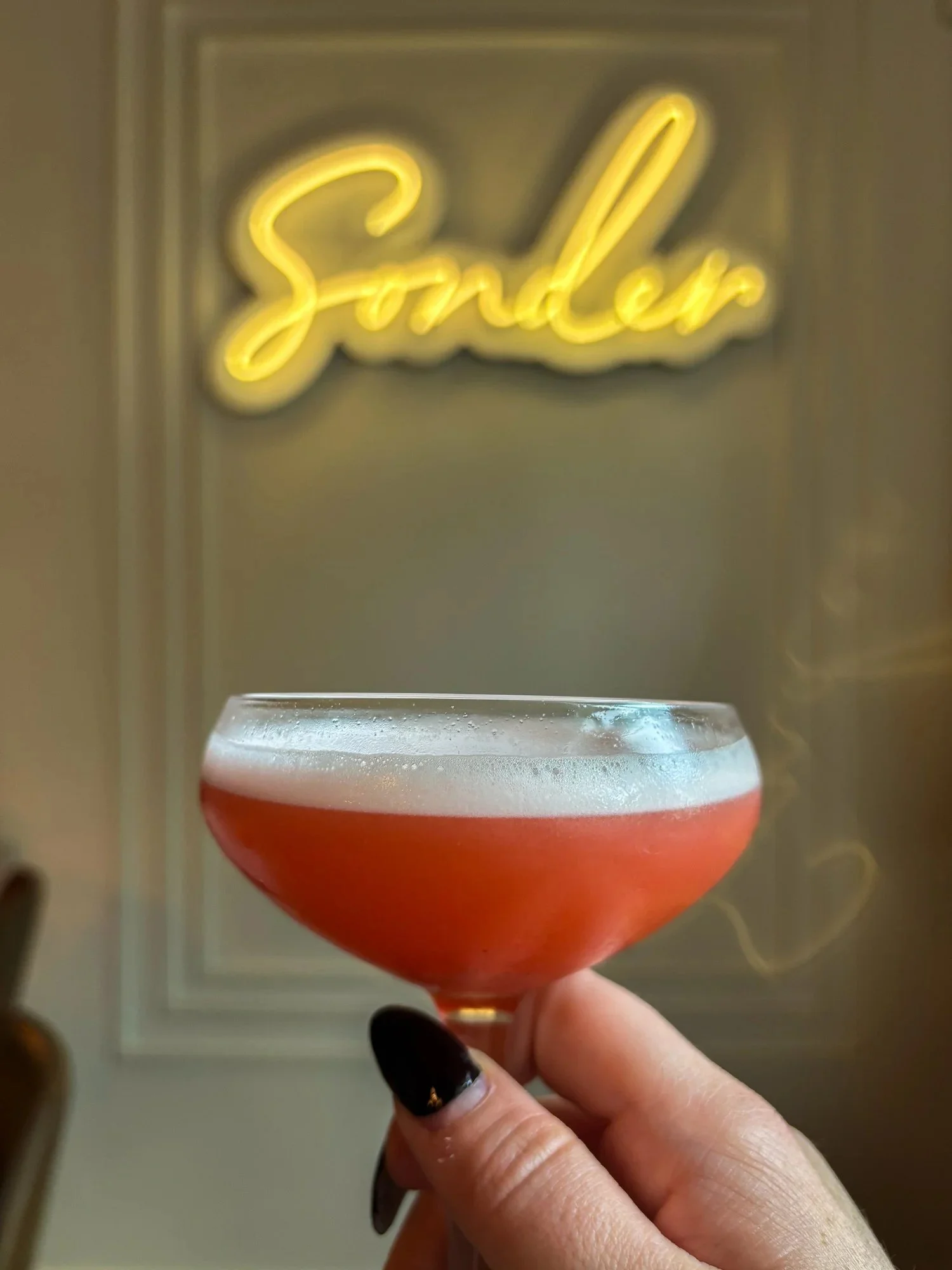 A hand holding a cocktail glass with a red drink and a foamy white top, seen in front of a yellow neon sign that says 'Saturday'.