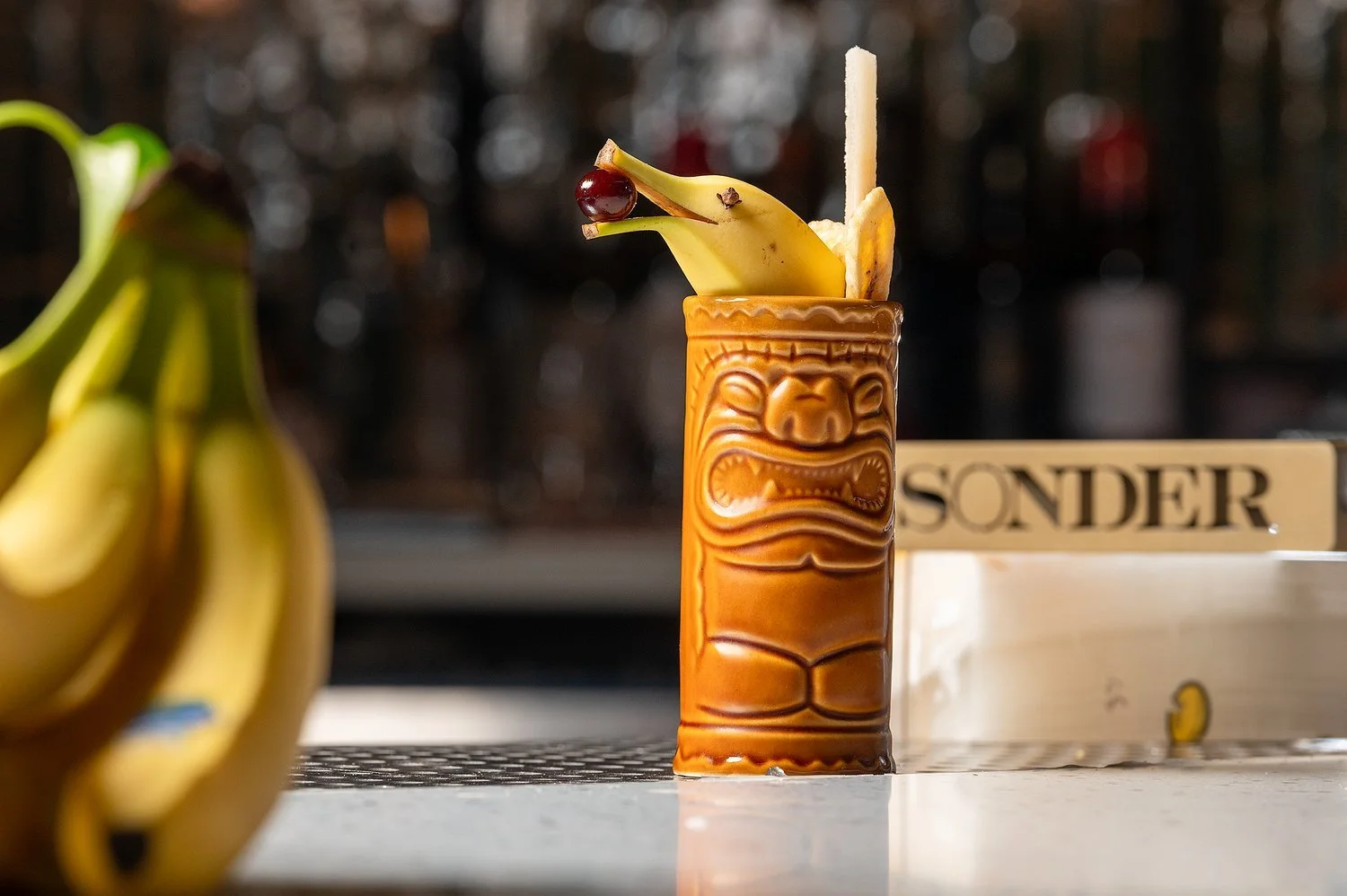 Hawaiian drink with a banana, cherry, and wafer in a tiki mug, with bananas on the left and a wooden sign with 'Sonder' in the background.