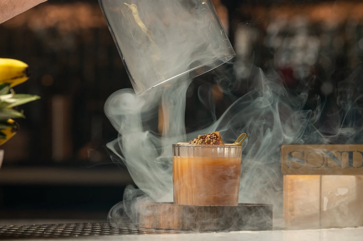 A glass of iced coffee or tea with steam and smoke rising, as a gloved hand pours more liquid from a clear glass container.