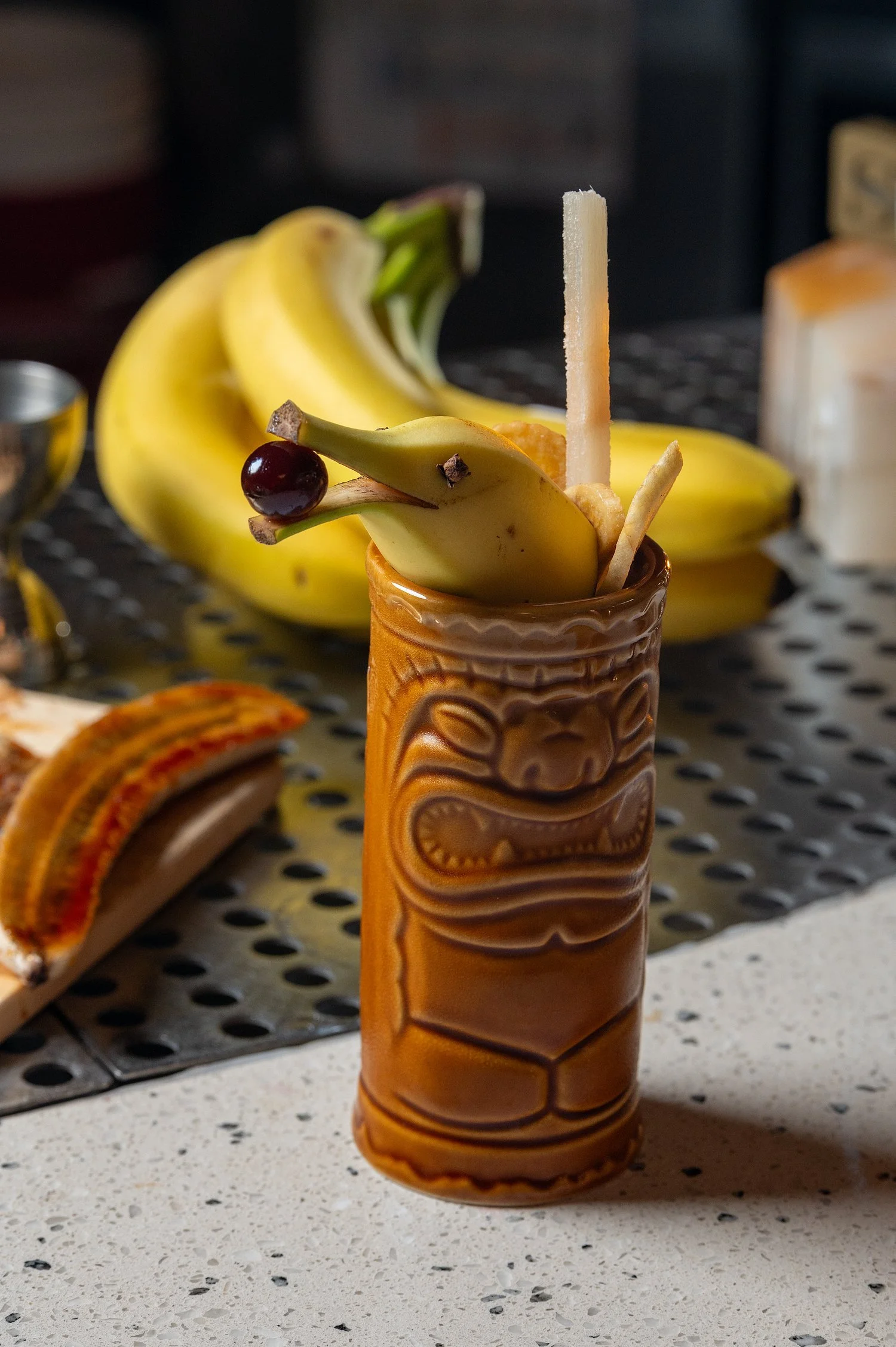 Traditional Hawaiian tiki mug with banana and cherry garnishes, placed on a white speckled countertop in front of a metal tray with baked goods.