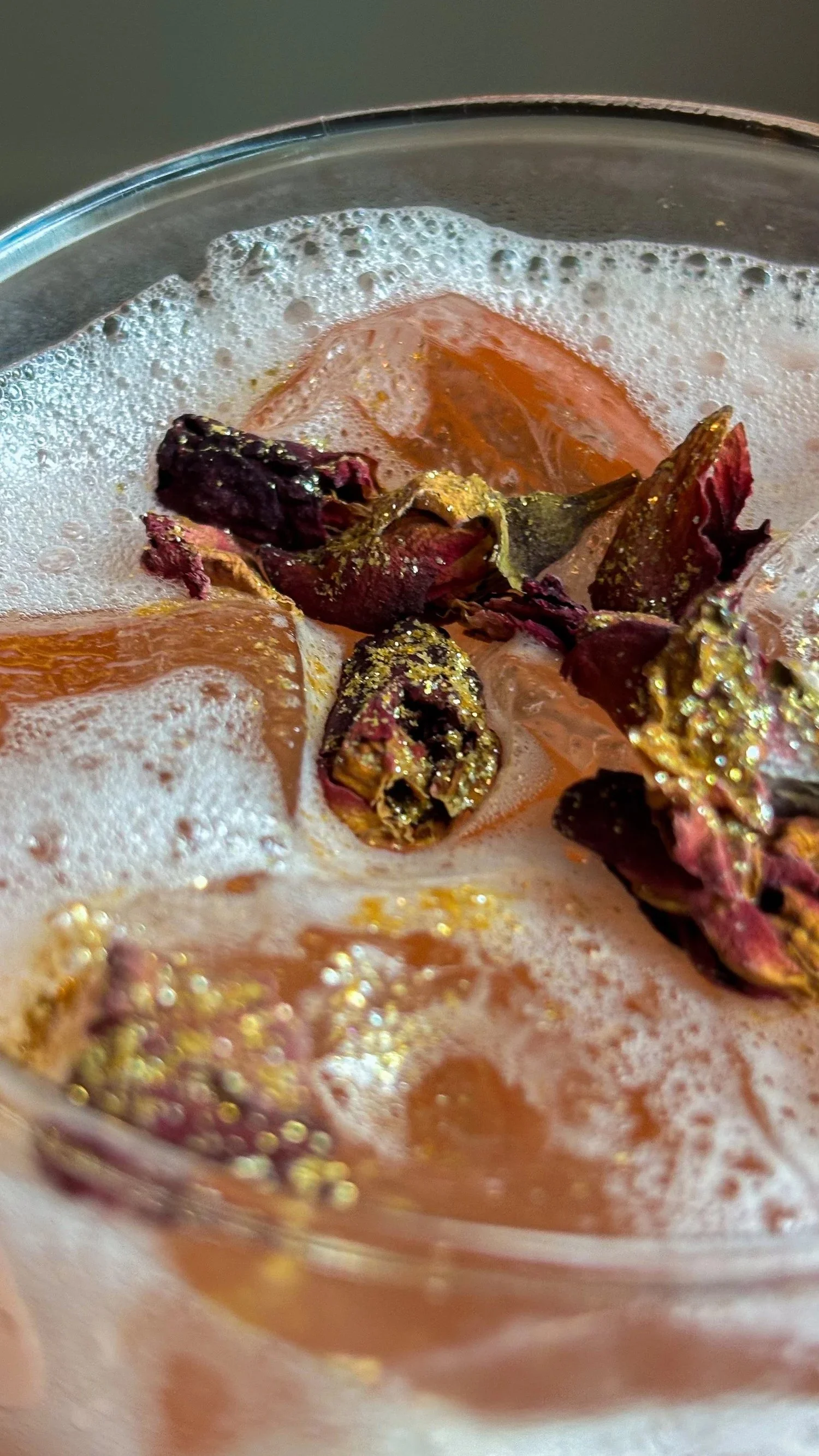 Close-up of a glass containing a beverage with foam, dried rose petals, and dried orange slices.