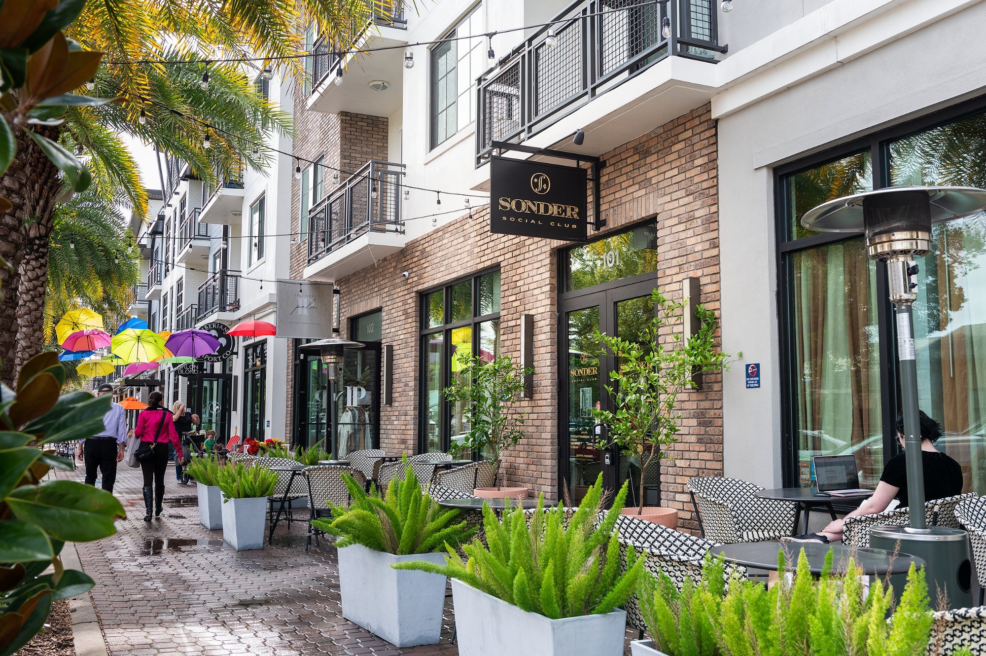 People walking on a sidewalk outside a modern building with a sign for Sonder Social Club. There are outdoor tables with chairs, potted plants, and decorative umbrellas. String lights are hanging above, with tall palm trees and greenery around.