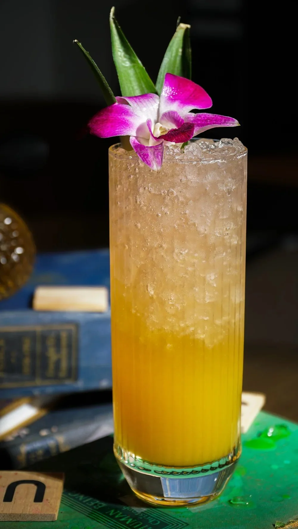 A tall glass of yellow-orange cocktail with crushed ice, garnished with a pink and white orchid flower and green pineapple leaves.