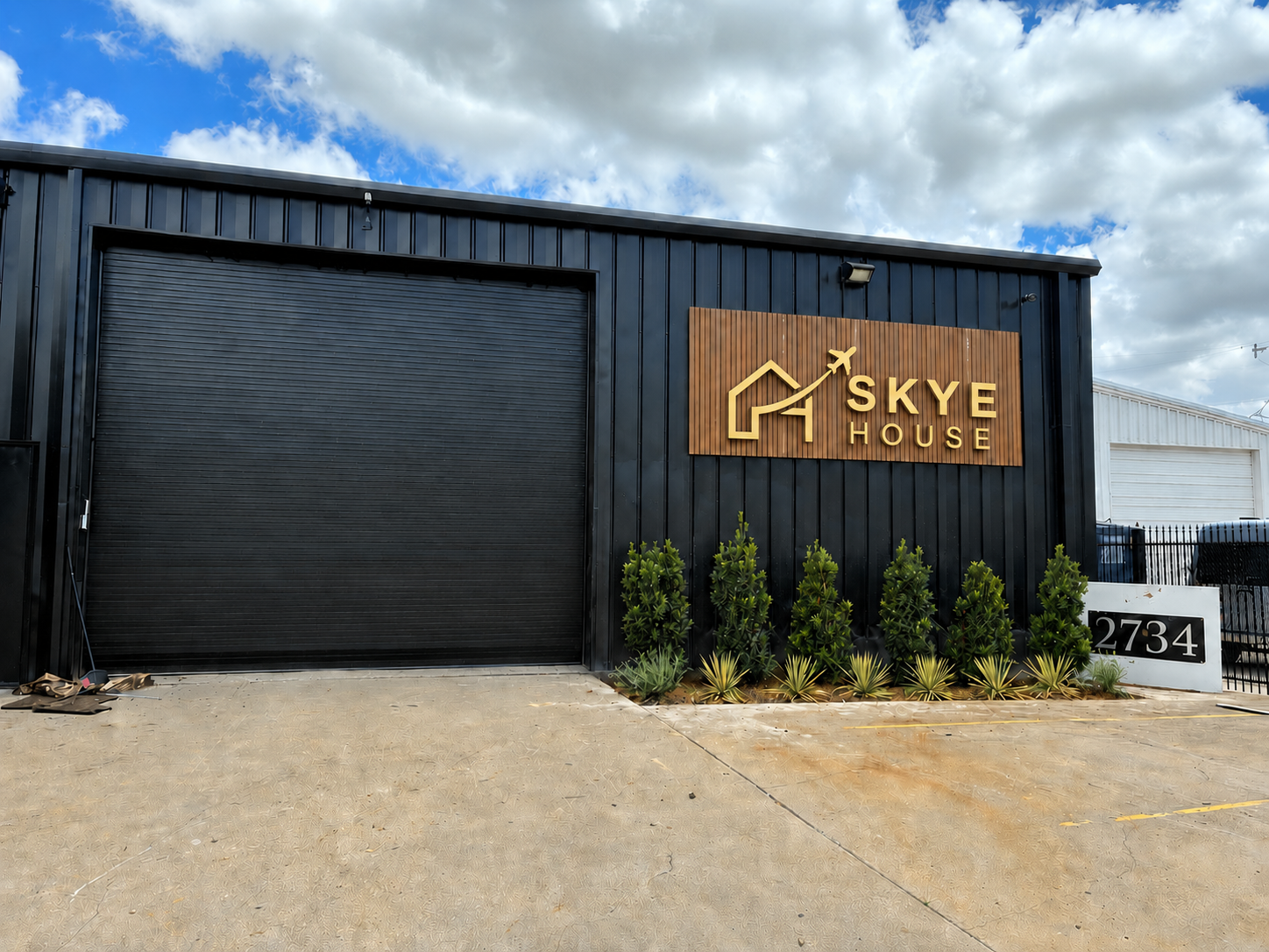Exterior of a building with a black metal wall, a large rolling black door, and a sign that reads "Skye House" with a gold house and upward arrow logo. The building address, 2734, is on a white sign. Small green plants are in front of the building, a