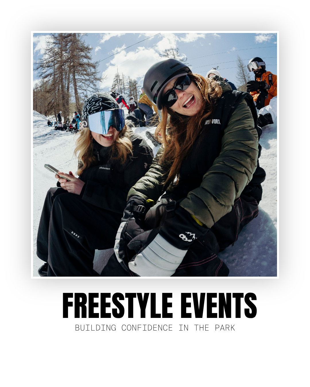 Two women wearing ski gear and helmets sitting on snow, smiling, with a group of skiers in the background and snow-covered trees under a partly cloudy sky.