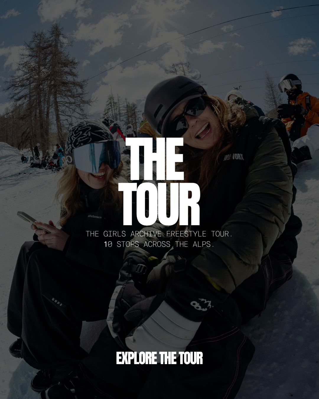 Women in winter gear and helmets smiling and taking a selfie on snow-covered mountains during a freestyle tour with other skiers in the background.