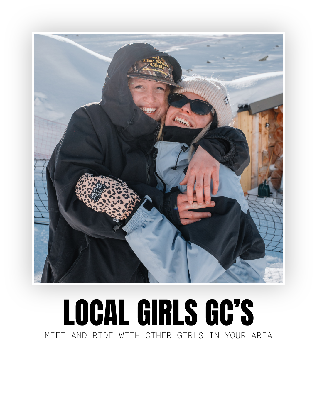 Two women in winter clothing hugging and smiling outdoors on a snowy day, with a wooden structure and snow-covered ground in the background.