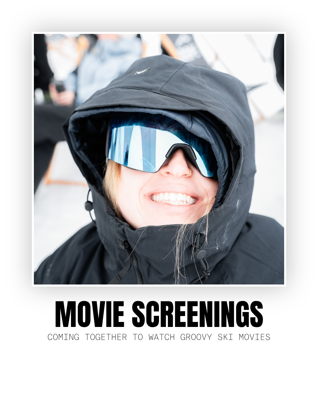 Person smiling with face partly covered by a large black hood and reflective ski goggles, wearing a black winter jacket, outdoors in snowy environment.