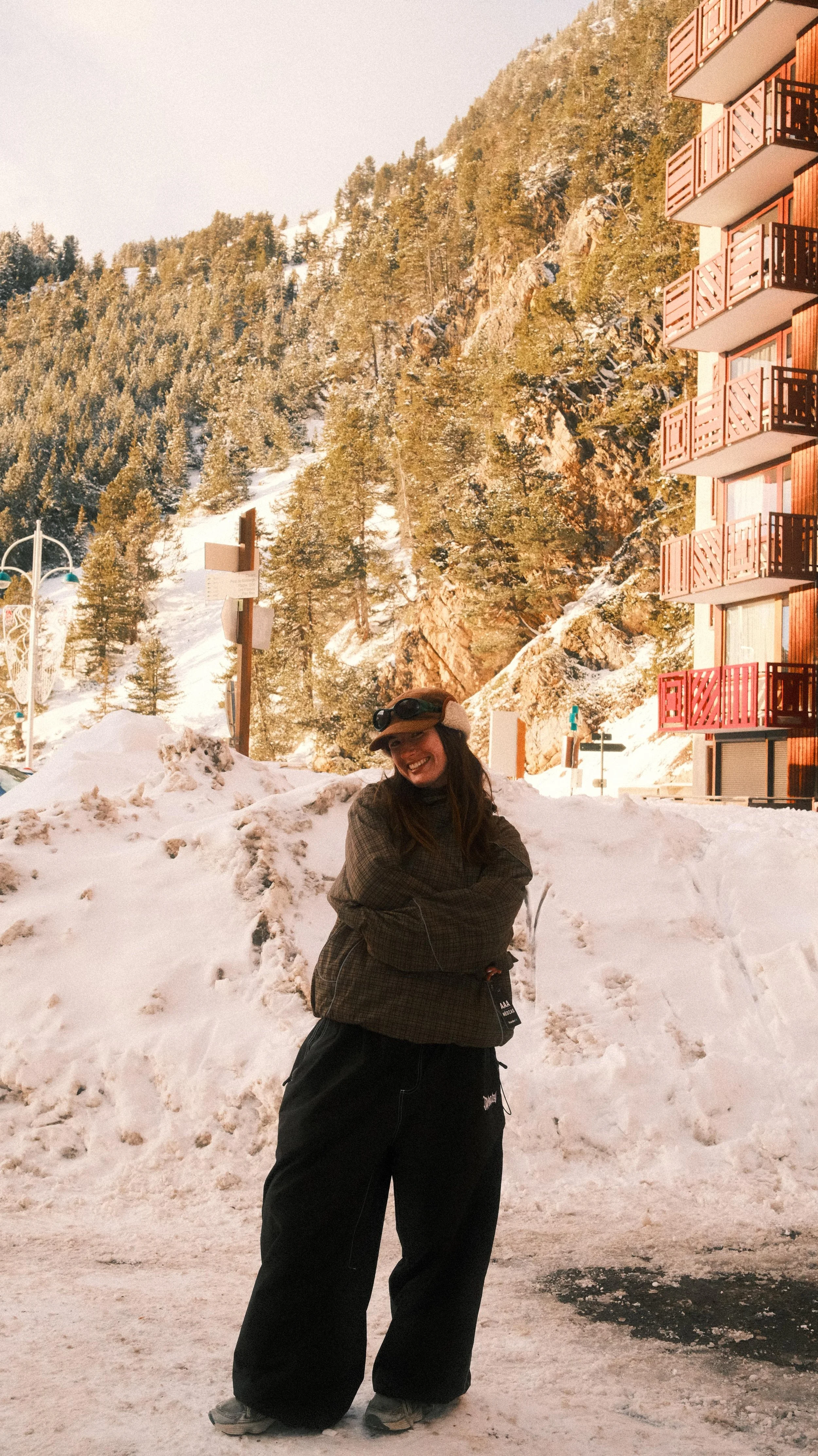 A smiling woman standing in snow with mountains and trees in the background, near a building with red balconies.