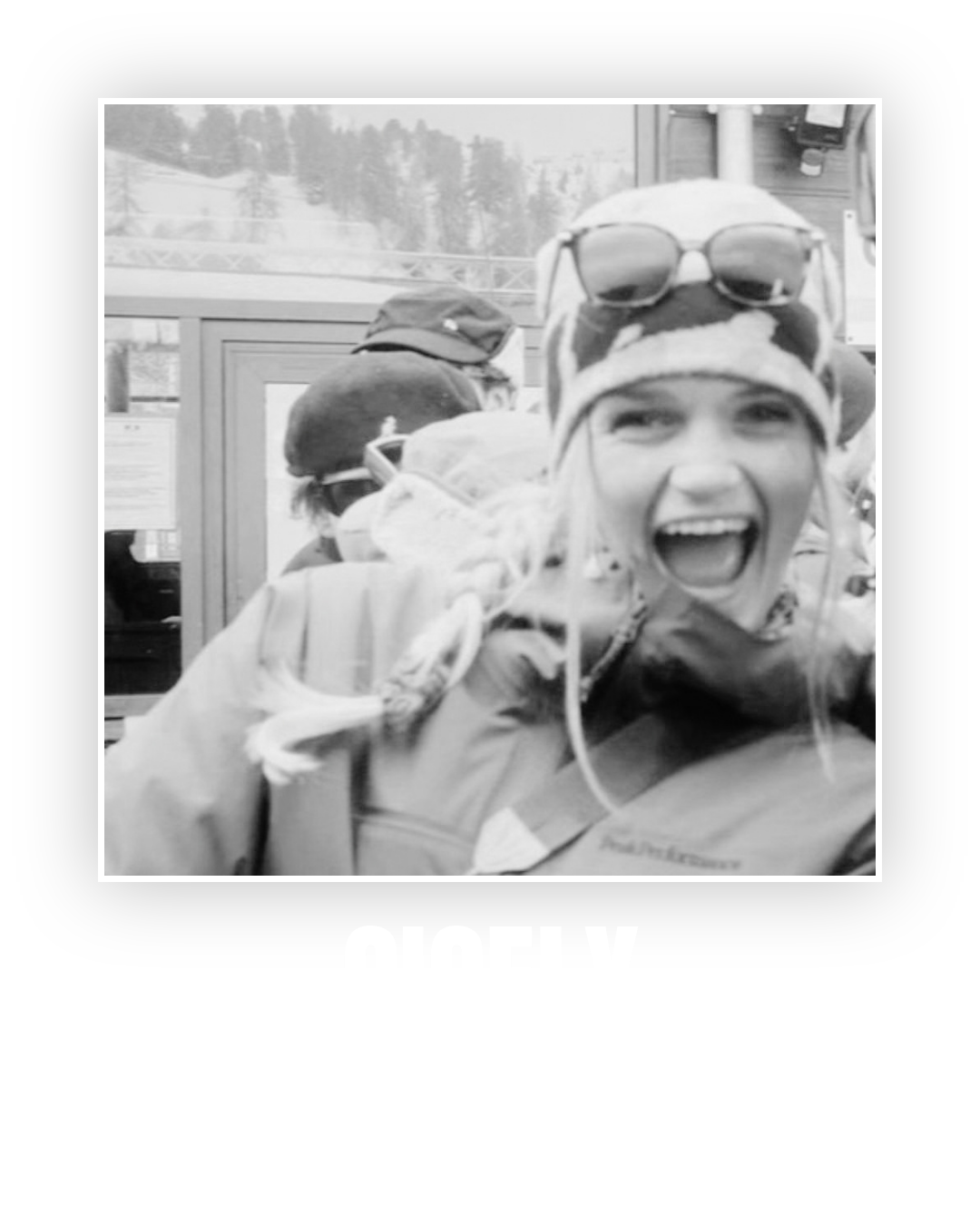 A black-and-white photo of a woman wearing outdoor gear, sunglasses on her head, and a patterned hat, smiling widely with her mouth open. The background shows other people and a mountainous landscape.