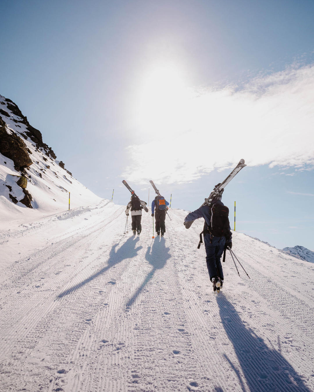 Three people hiking up a snow-covered mountain trail with skis on their shoulders under a bright sun.
