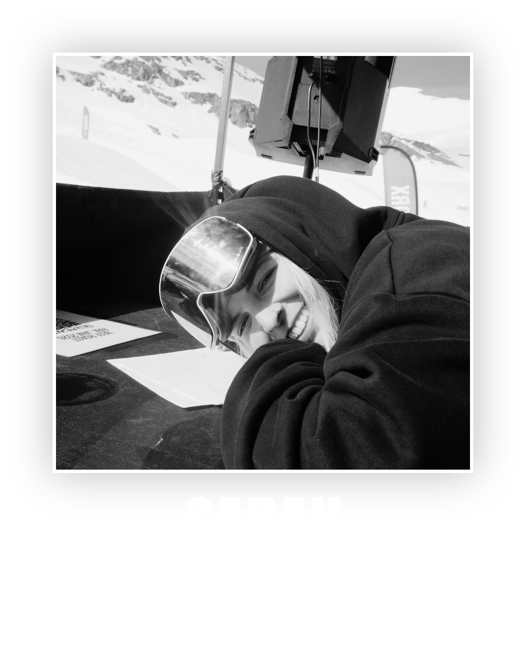 A woman in ski goggles and a black hoodie smiling and resting her head on her arms on a table at a snowy mountain ski resort.