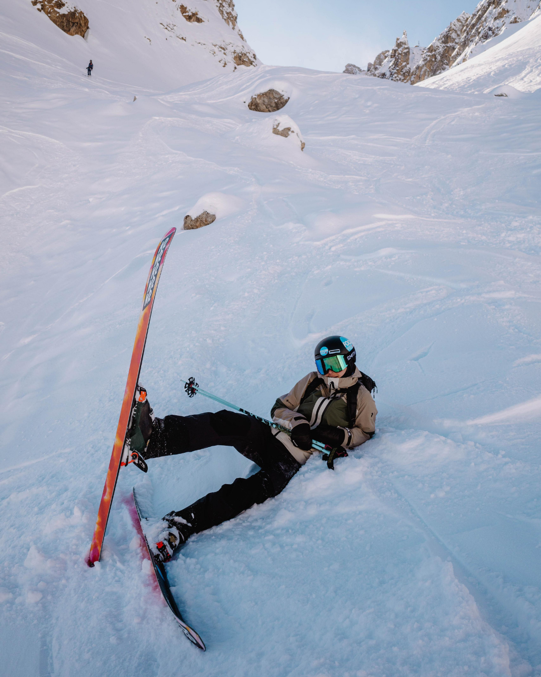 A skier has fallen and is lying on the snow with skis and poles. The skier is wearing a helmet, goggles, and winter gear in a snowy mountain landscape.
