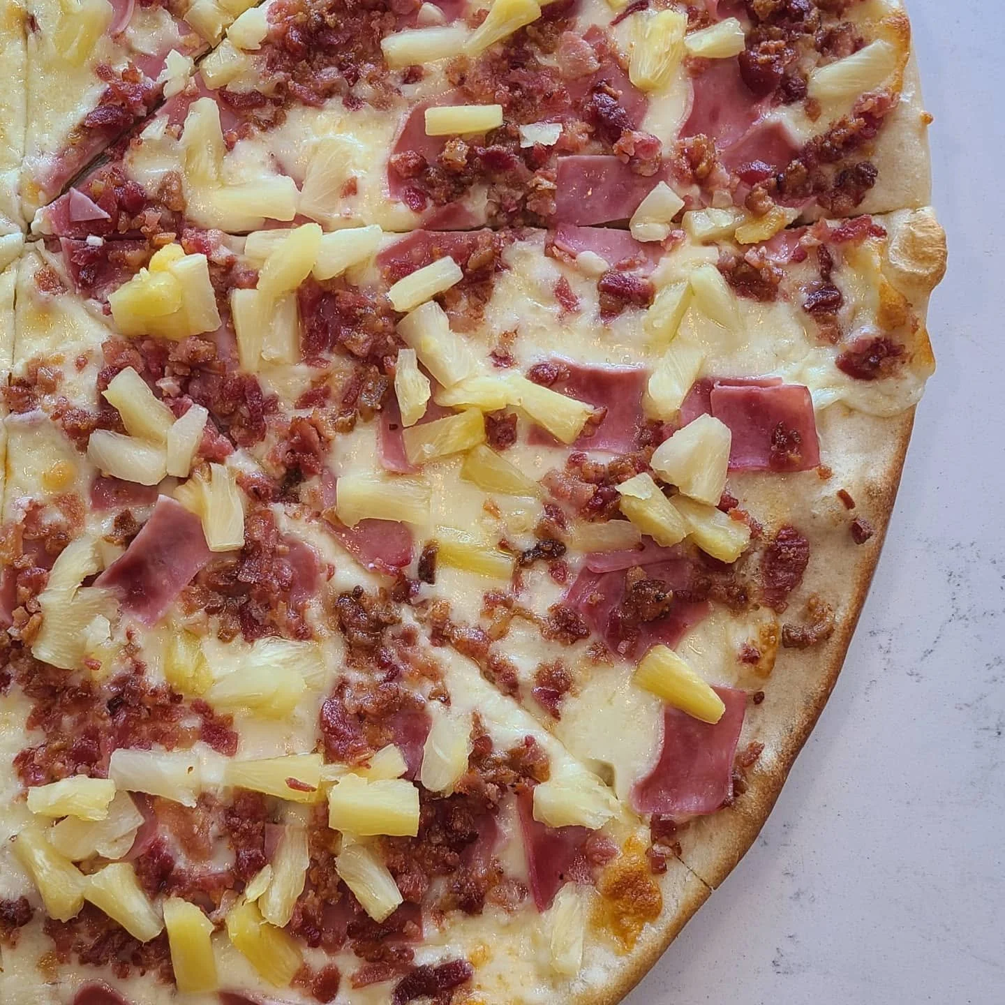 Close-up of a Hawaiian pizza topped with cheese, ham, bacon, ground meat, and pineapple on a thin crust.