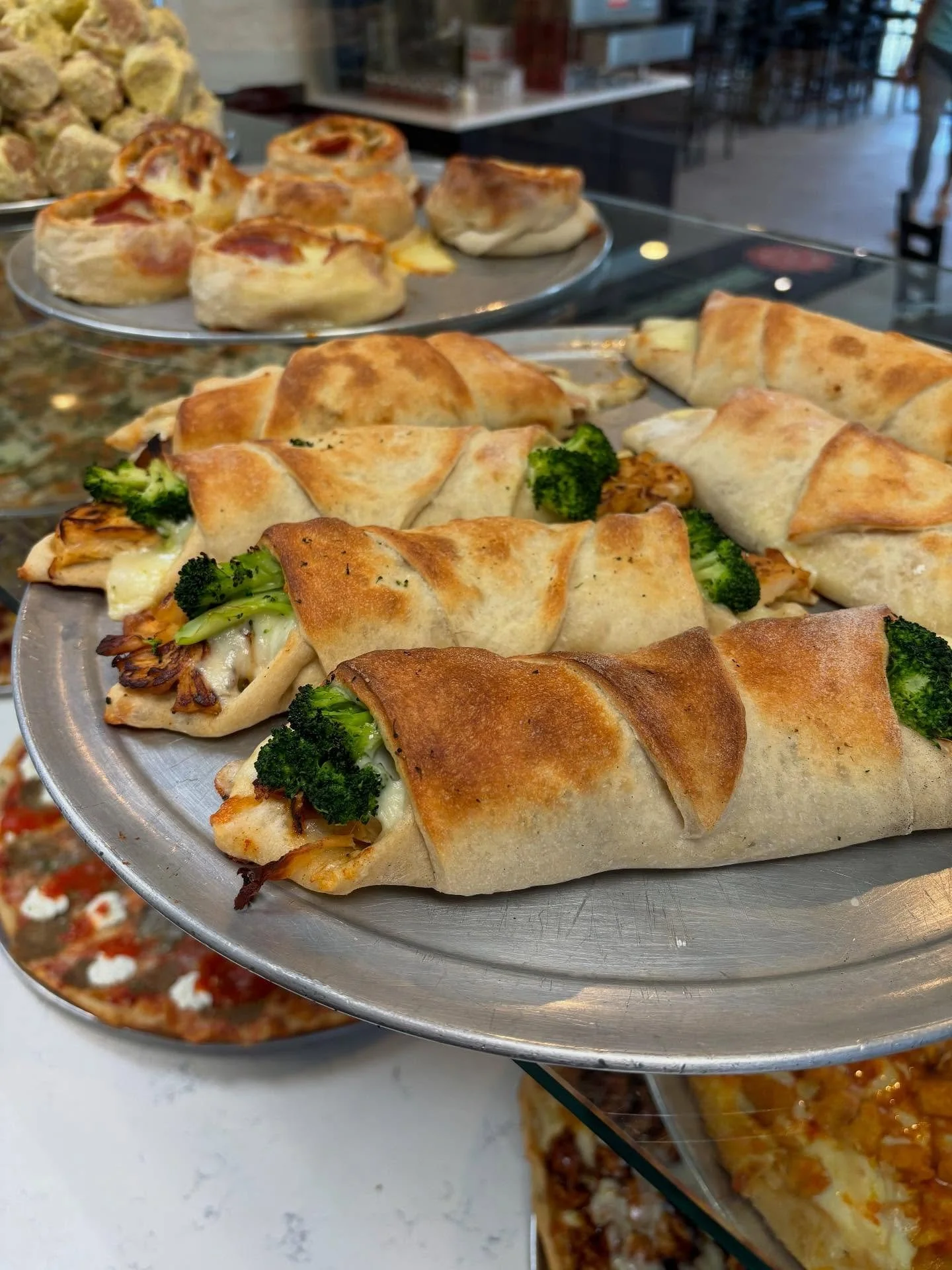 Plate of baked chicken and broccoli wrapped in flatbread, stacked with additional baked items in the background at a restaurant or bakery.