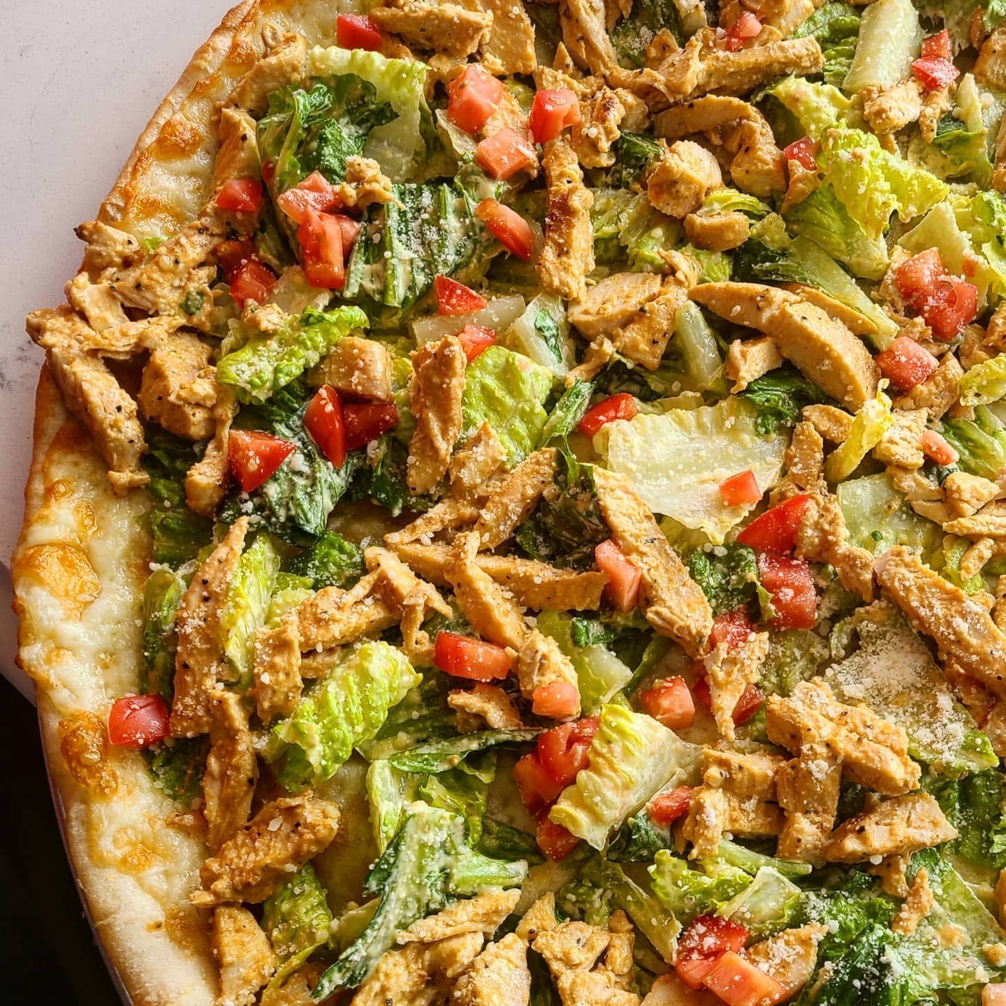 Close-up of a pizza topped with shredded chicken, chopped lettuce, diced tomatoes, and shredded cheese, with a golden crust.