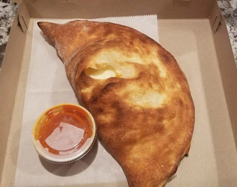 A calzone pizza in a box with a small cup of marinara sauce on the side.