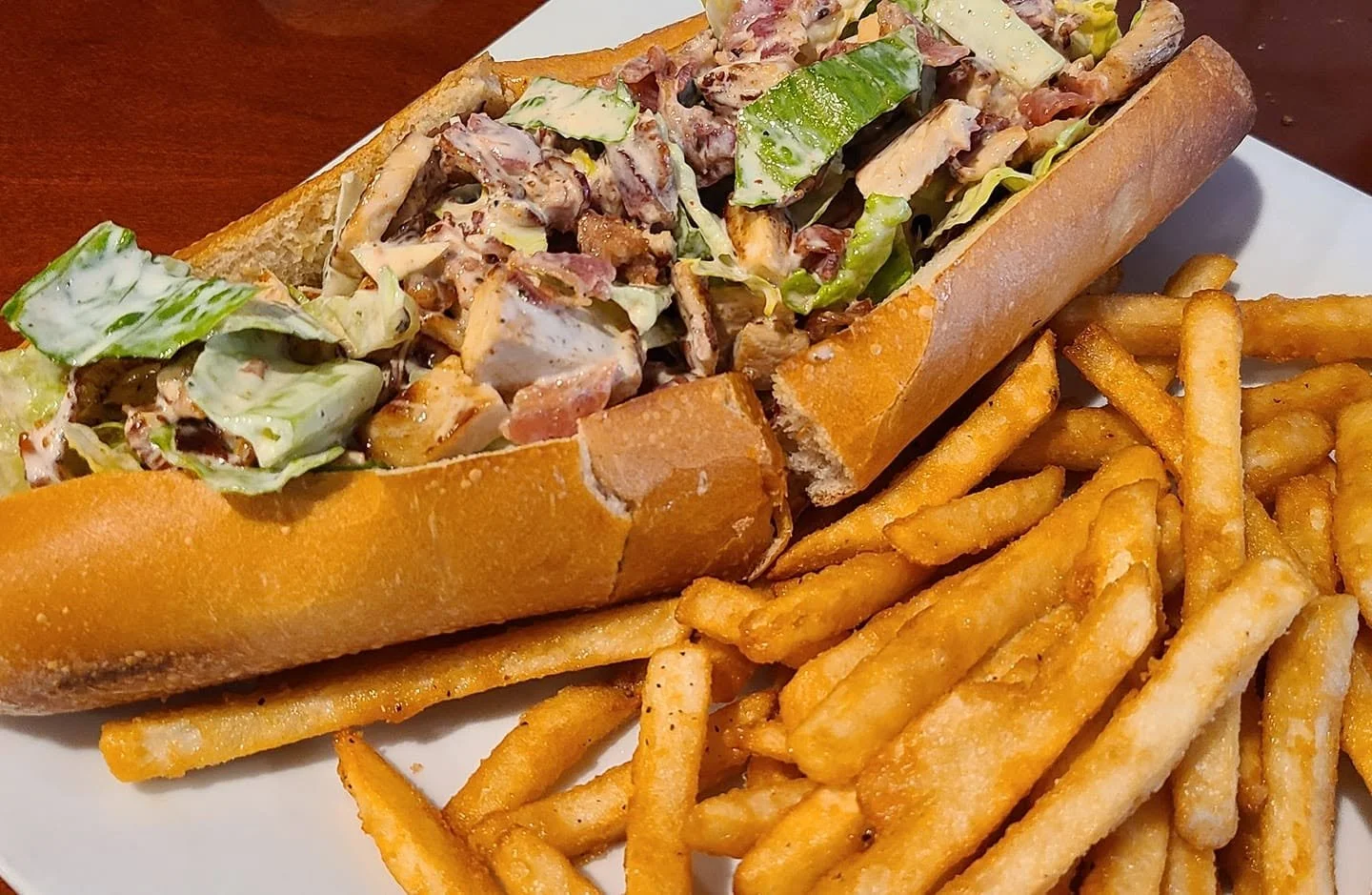 Sub sandwich with lettuce, bacon, and creamy dressing on a hoagie roll, served with a side of crinkle-cut French fries on a white plate.