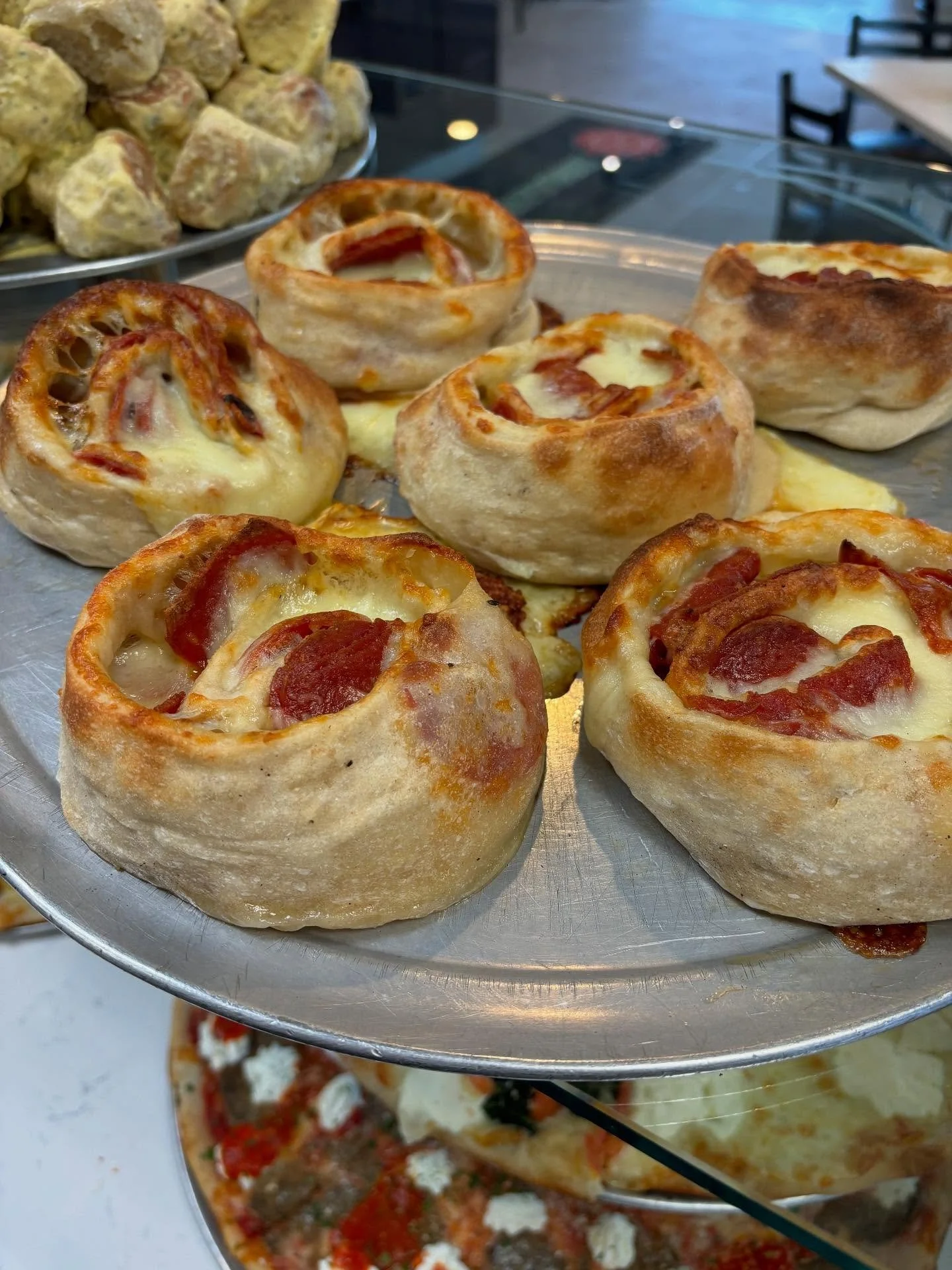 Close-up of a metal tray filled with small, round pizza bread rolls topped with cheese and pepperoni slices, with a second tray of similar rolls and a tray of cousin snacks visible in the background.