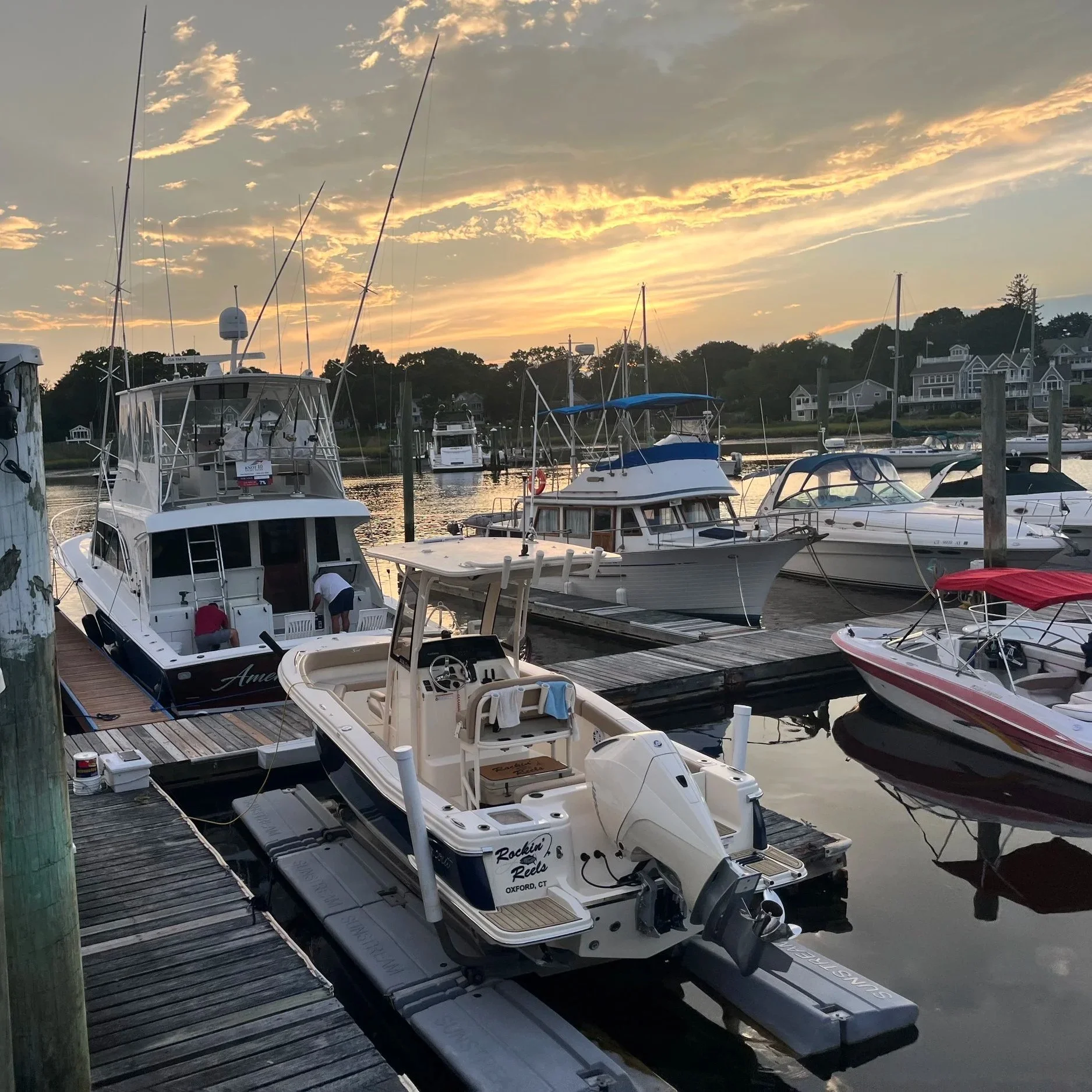 Boats docked at a marina during sunset with houses on a hillside in the background.