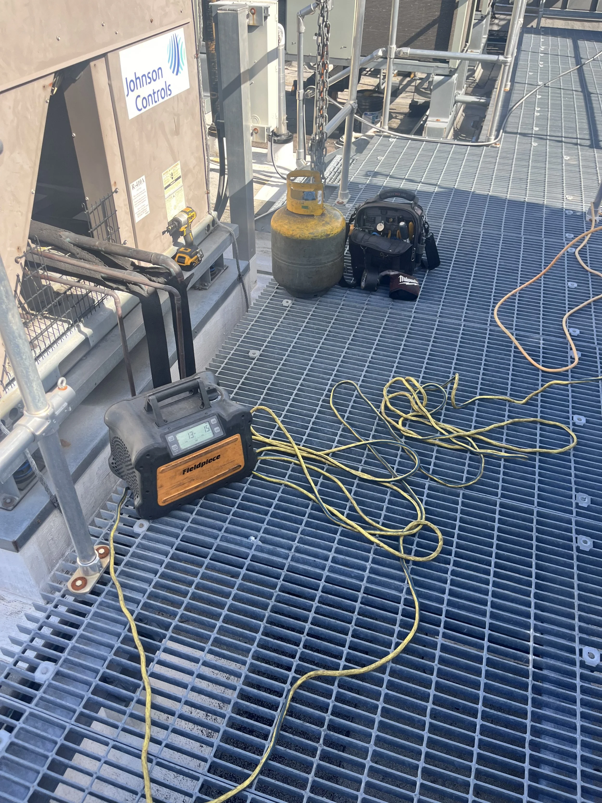 Industrial equipment on grated flooring with tools, a gas cylinder hanging from a chain, a digital device, and electrical cords, likely on a rooftop or maintenance platform.