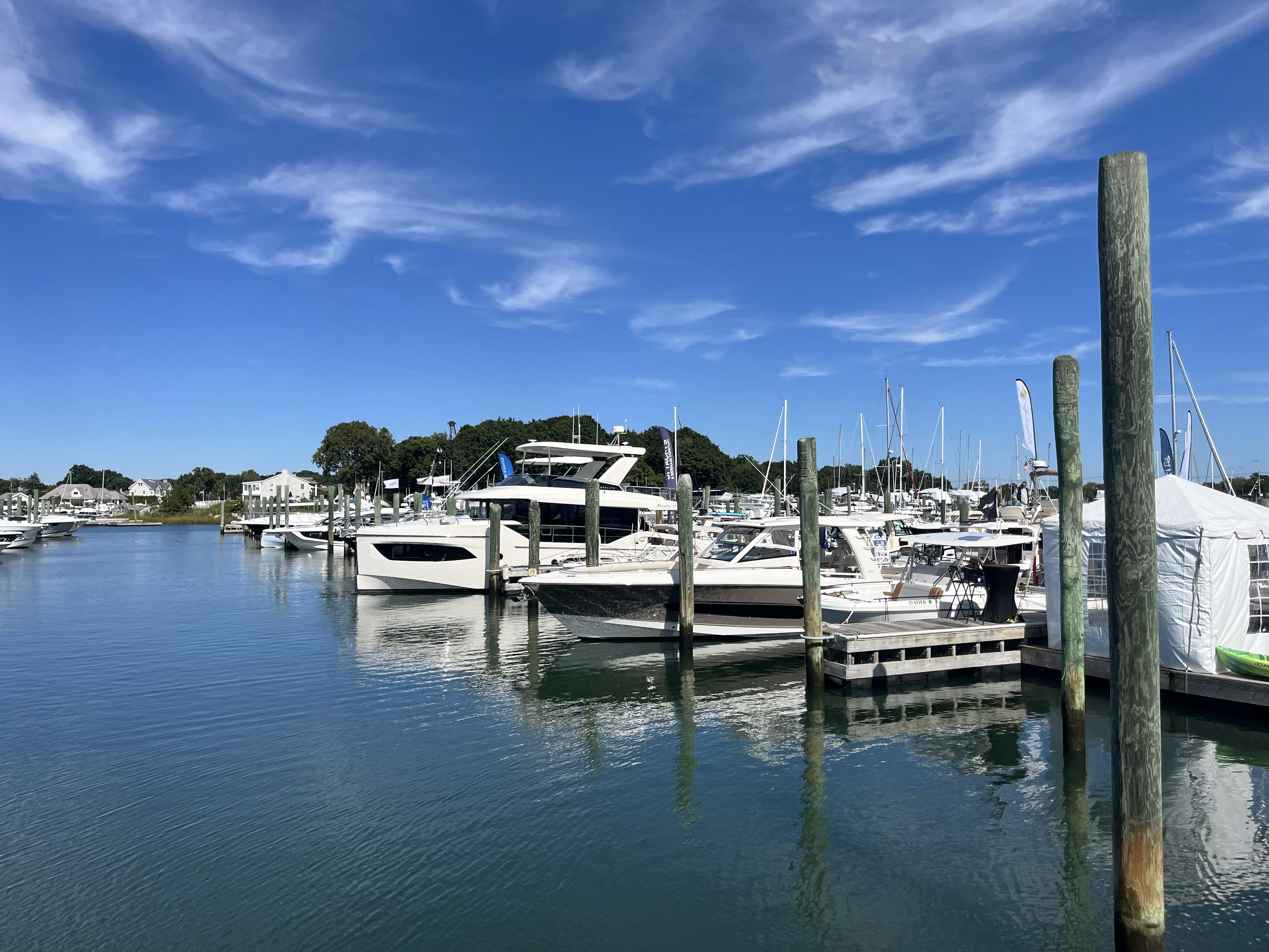 A marina with boats docked, wooden posts, calm water, houses on the shore, and a bright blue sky with wispy clouds.