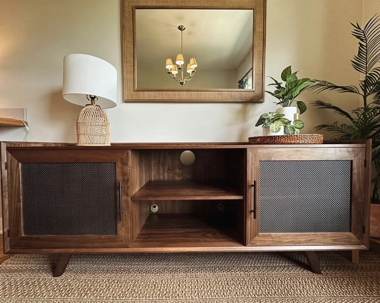 A mid-century modern wooden sideboard with open and closed storage compartments, decorated with a table lamp, potted plants, and a large wall mirror, in a room with beige walls and a woven rug.