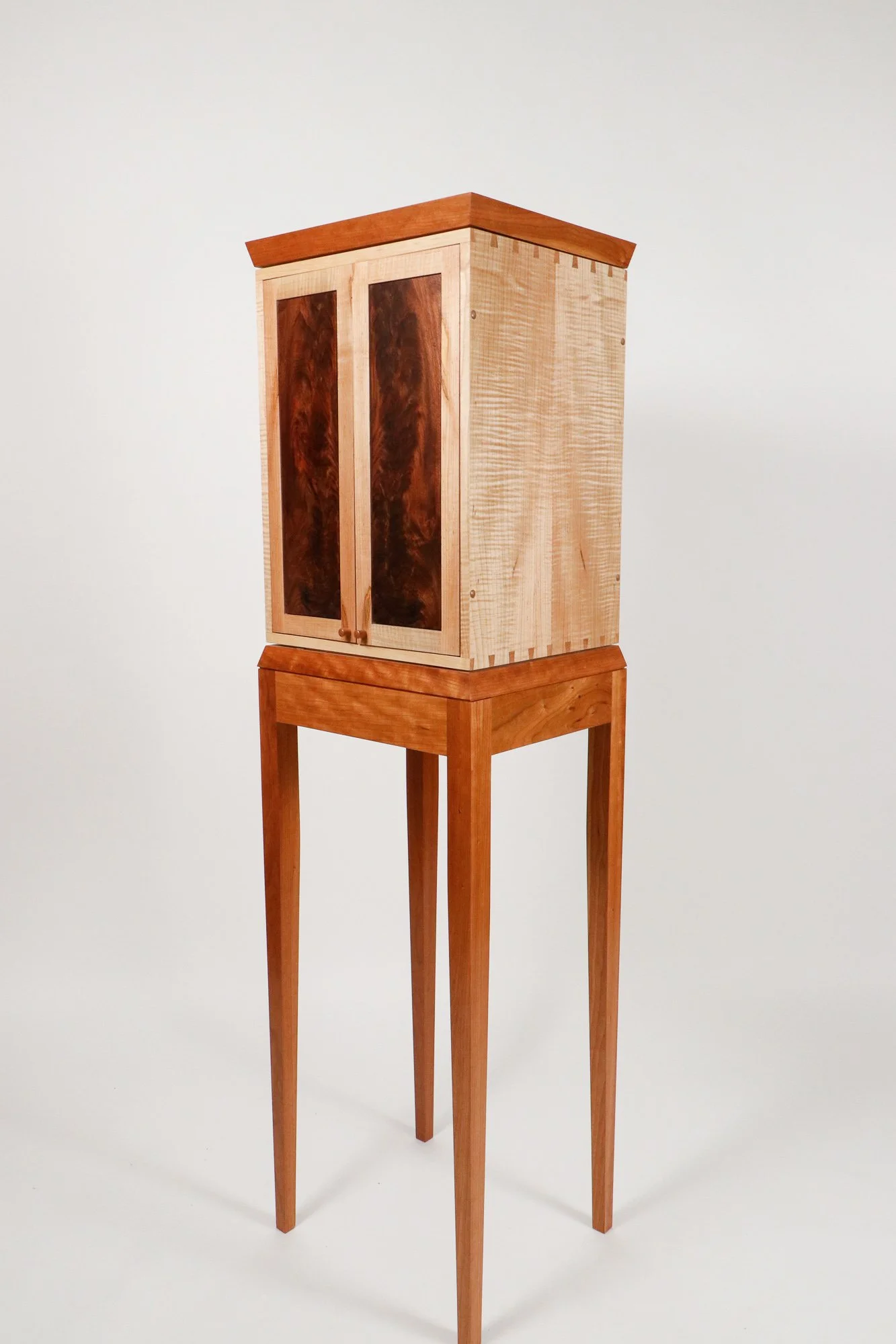A handcrafted wooden cabinet on tall, thin legs, with two doors featuring darker wood panels, set against a plain white background.