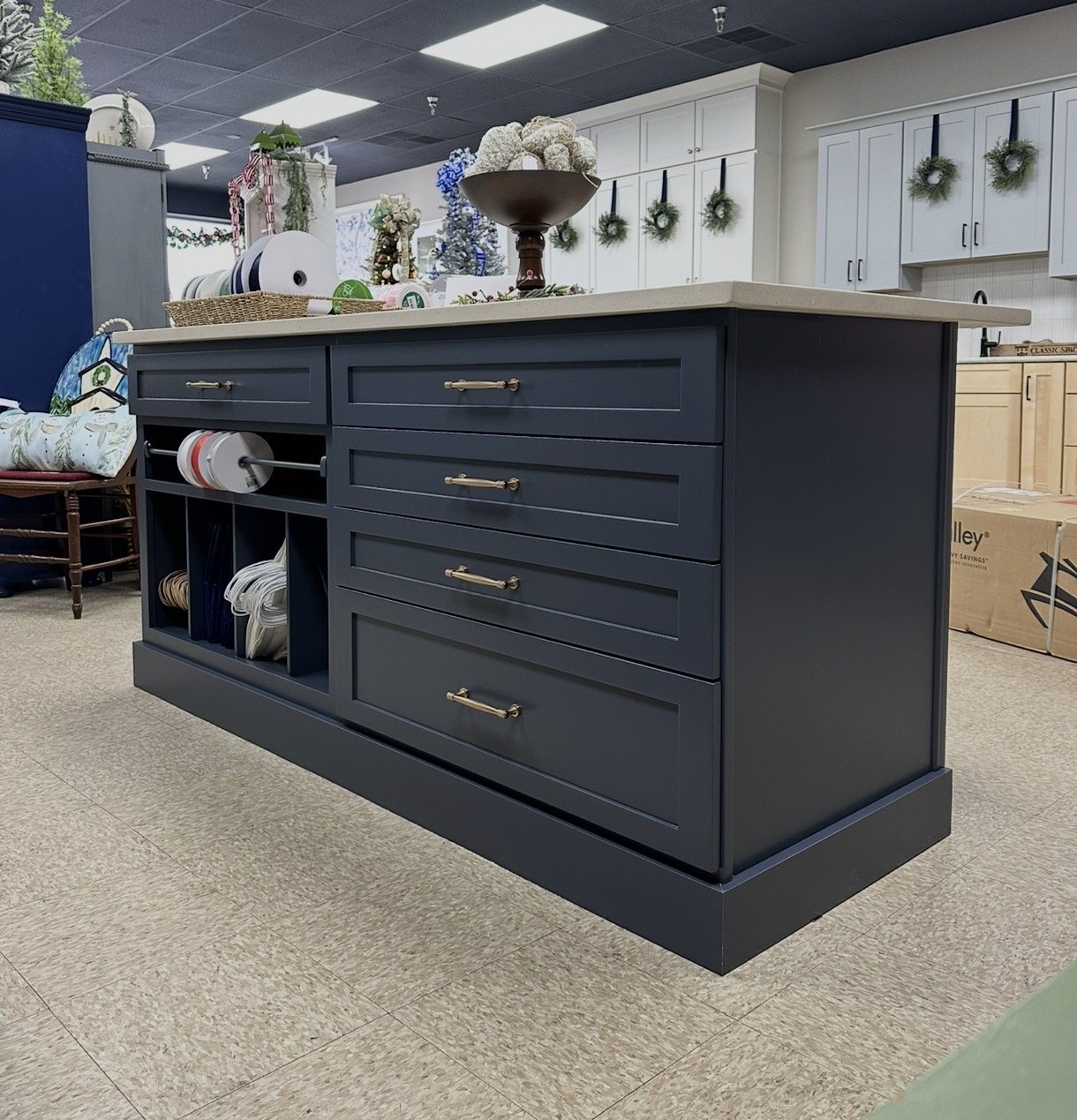 A navy blue kitchen island with a white countertop, featuring drawers, cabinets, and open shelves with rolls of wire, and decorative items on top, in a store decorated for Christmas.