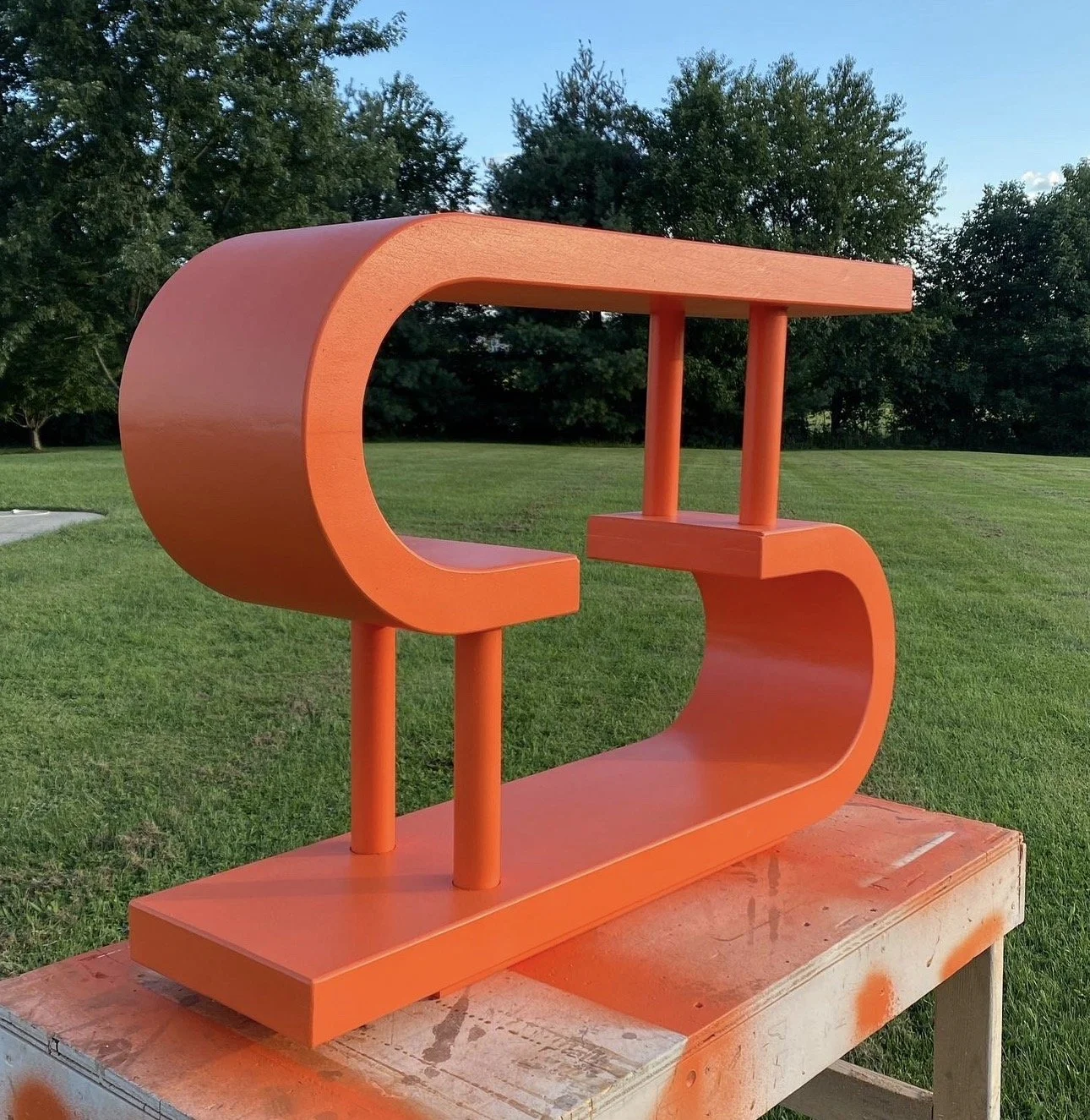Orange modern sculpture outdoors on a grassy field with trees in the background.