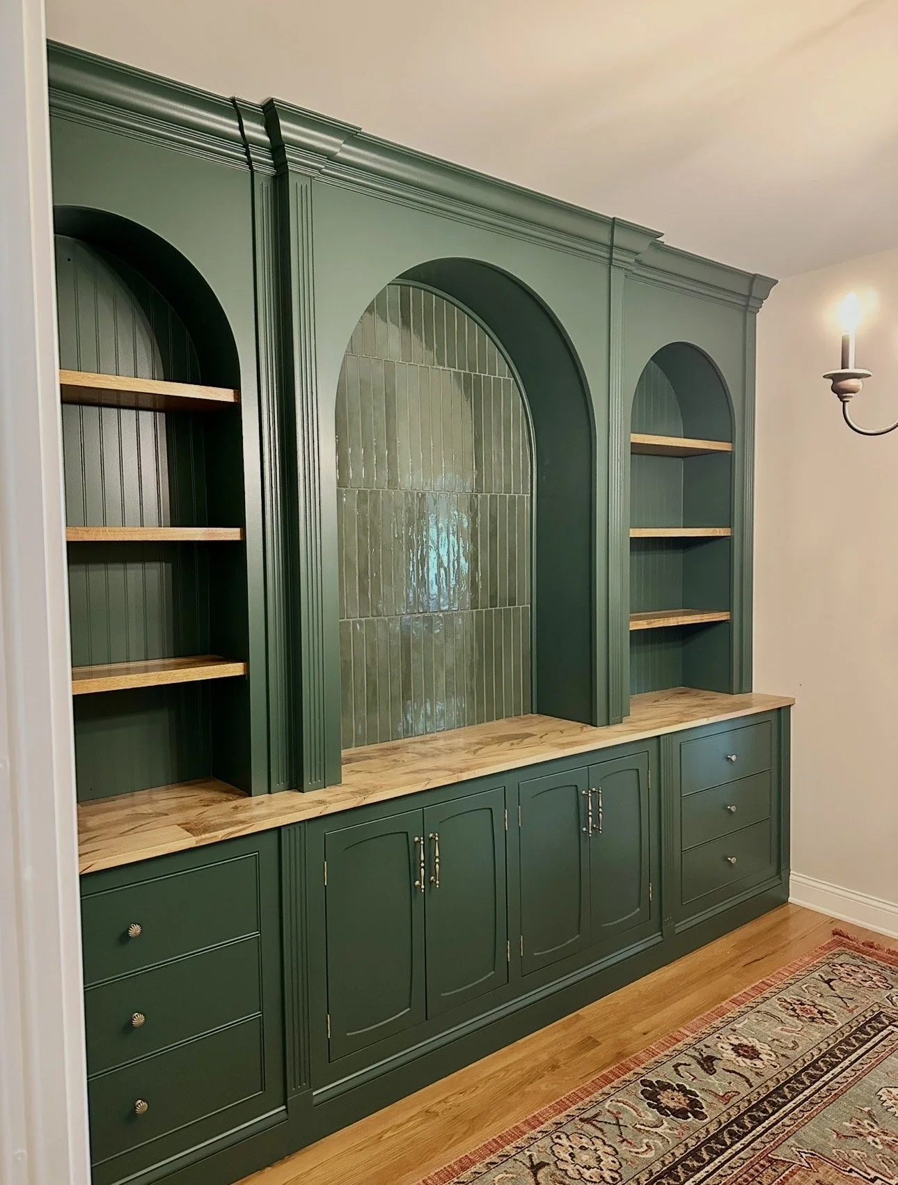 Green built-in bookshelf with wooden shelves, decorative arch design, and closed cabinets below, in a living room with a patterned rug and wall sconce lighting.