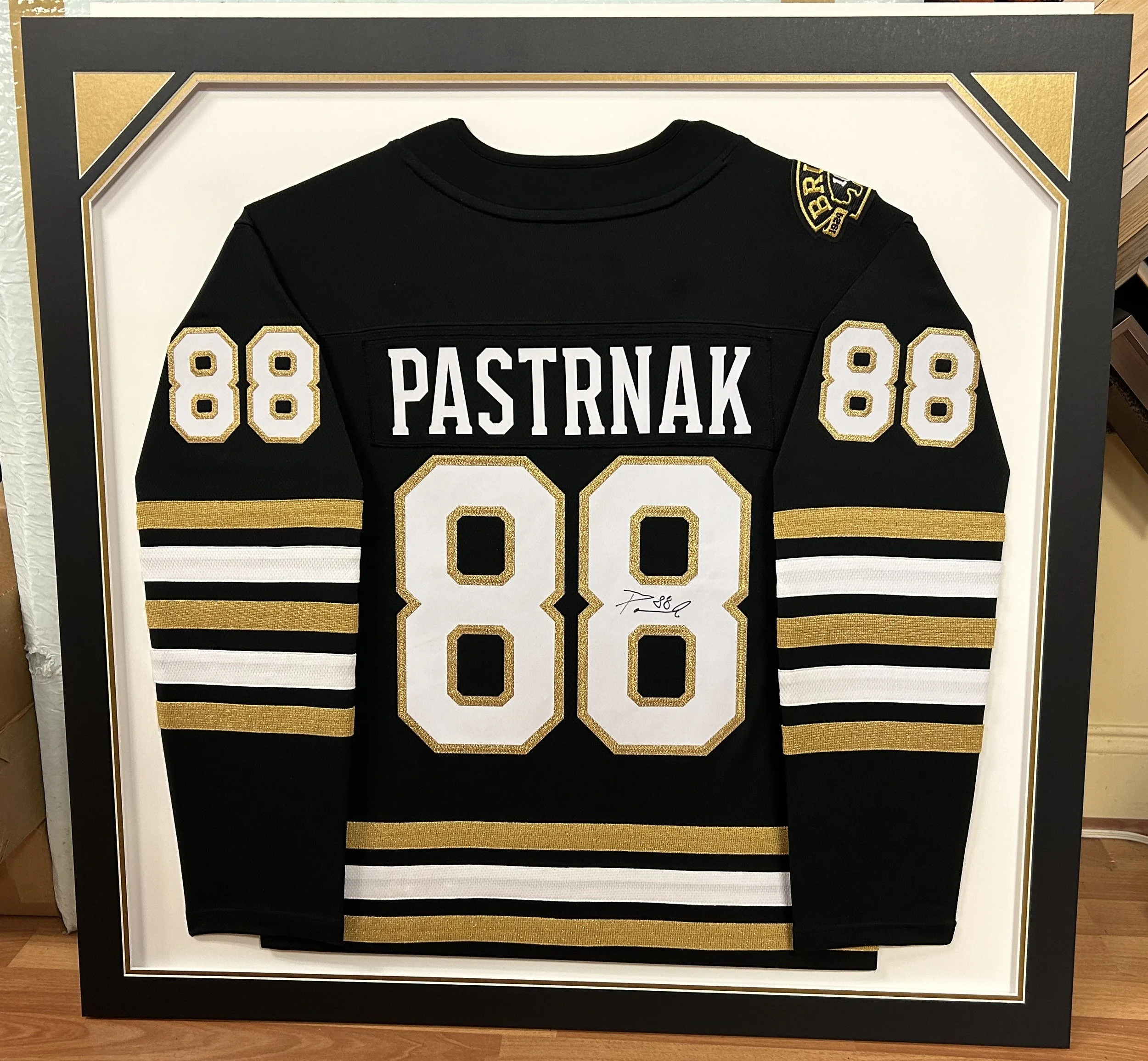Autographed black and gold Boston Bruins number 88 Pastrnak hockey jersey, custom framed in a black shadow box with a white and gold mat. Custom framing by Caran Art & Frame.