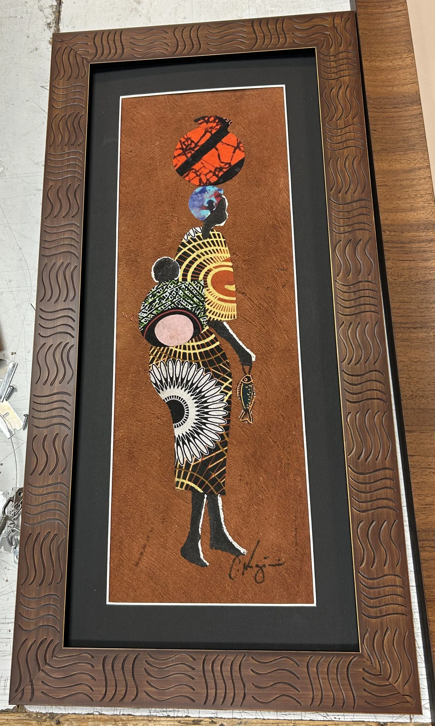 Tall artwork of an African woman carrying a basket on her head and a baby on her back, in a carved wavy-pattern brown wood frame with a black mat. Custom framing by Caran Art & Frame.