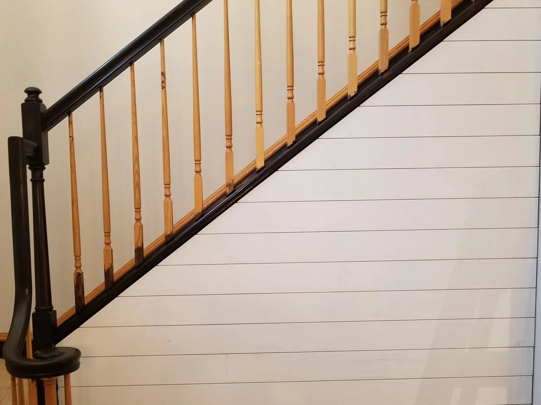 Interior staircase with light-colored wooden handrail and balusters, black trim, and white horizontal wall paneling.