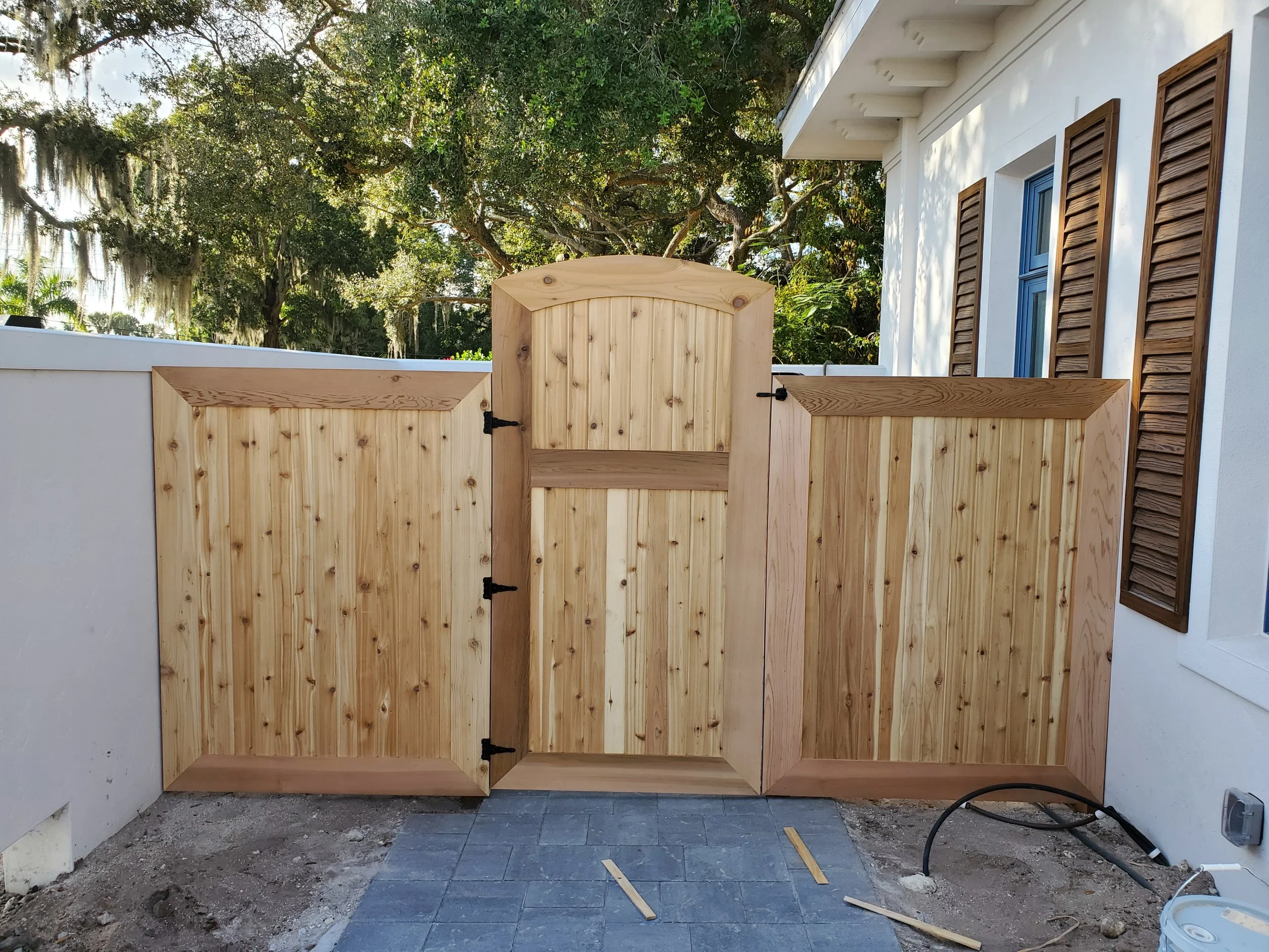 Wooden gate attached to a white wall outside a house with brown shutters, with trees and greenery in the background during daytime.