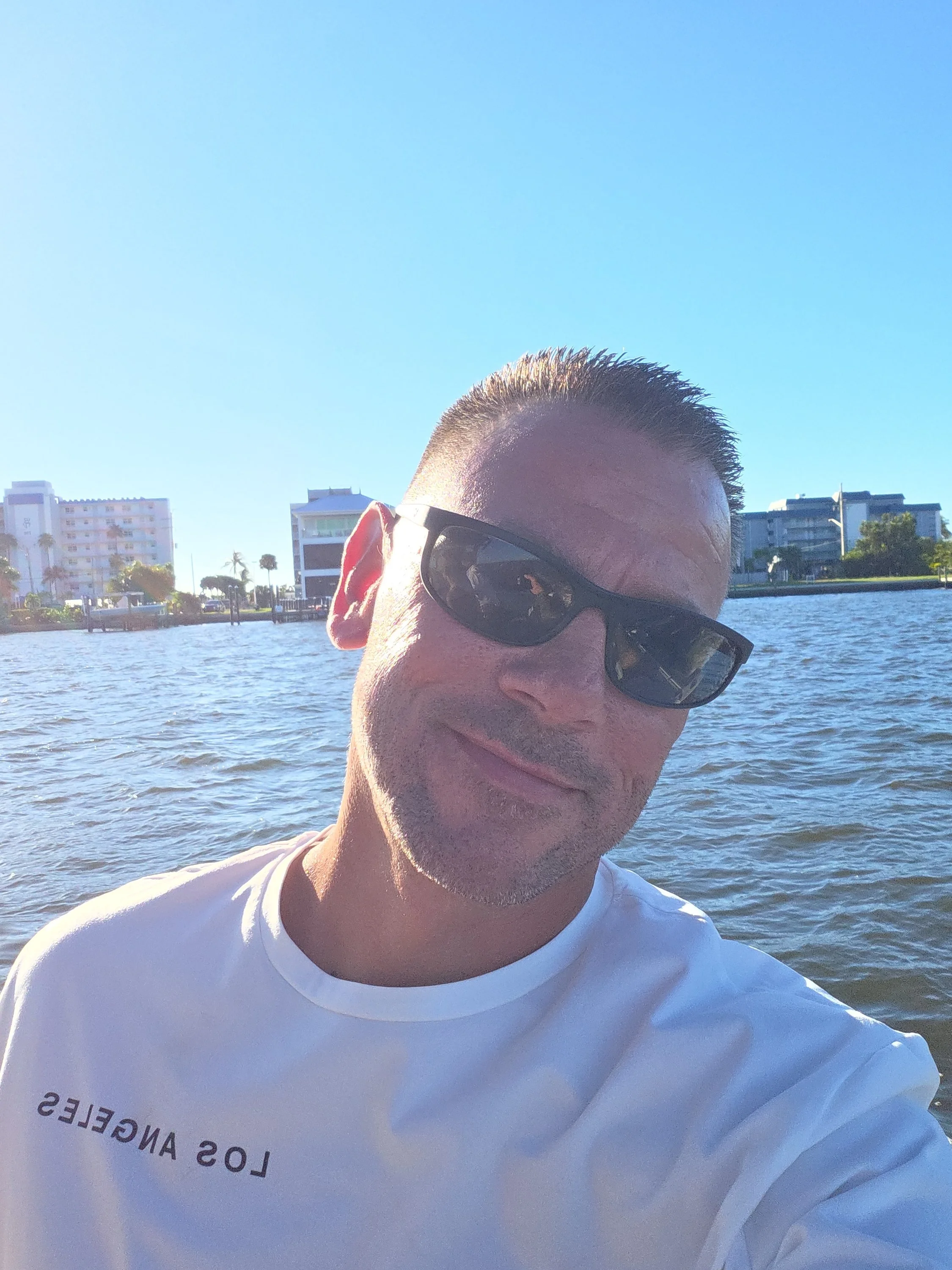 A man with short hair, wearing sunglasses and a white shirt, standing outdoors near water with buildings and trees in the background.