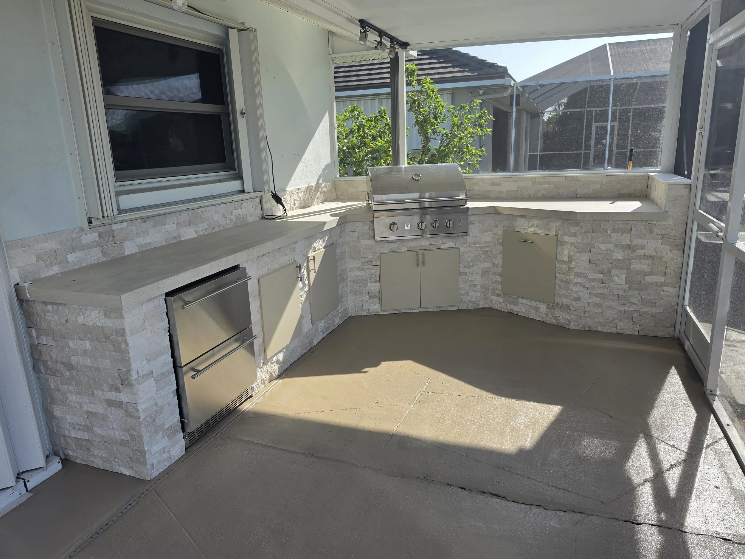 A screened-in patio area with a built-in barbecue grill, beige stone wall, and large sliding window with black screens. There are cabinets below the grill and a small window above it, with outdoors visible through the screens.