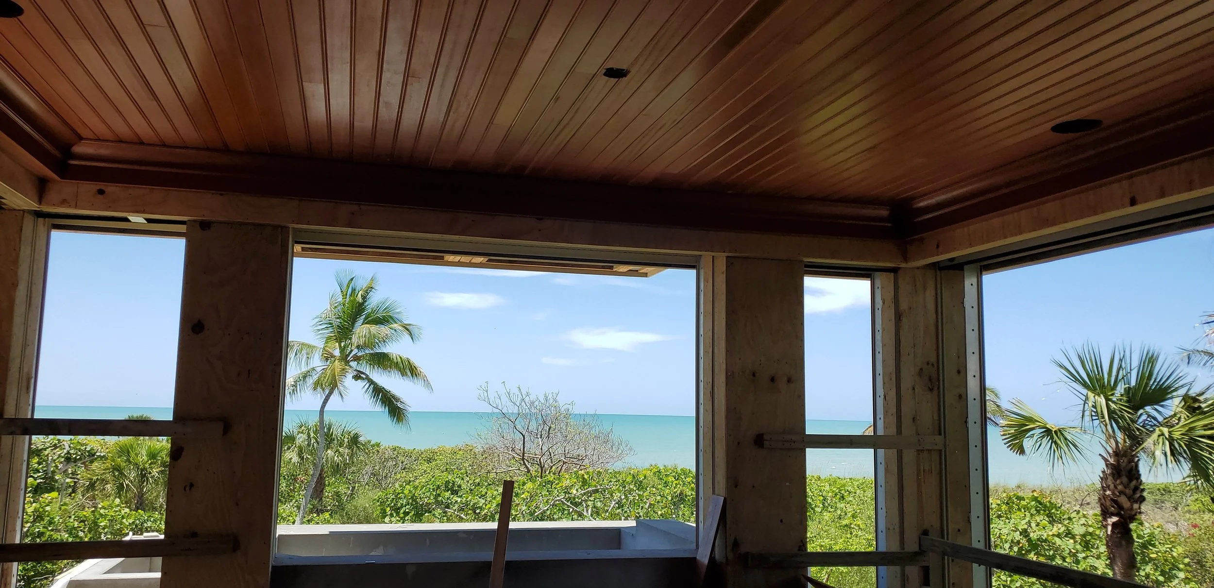 Interior view of a building under construction with framed walls and a wooden ceiling, overlooking a tropical landscape with palm trees, bushes, and the ocean in the distance.