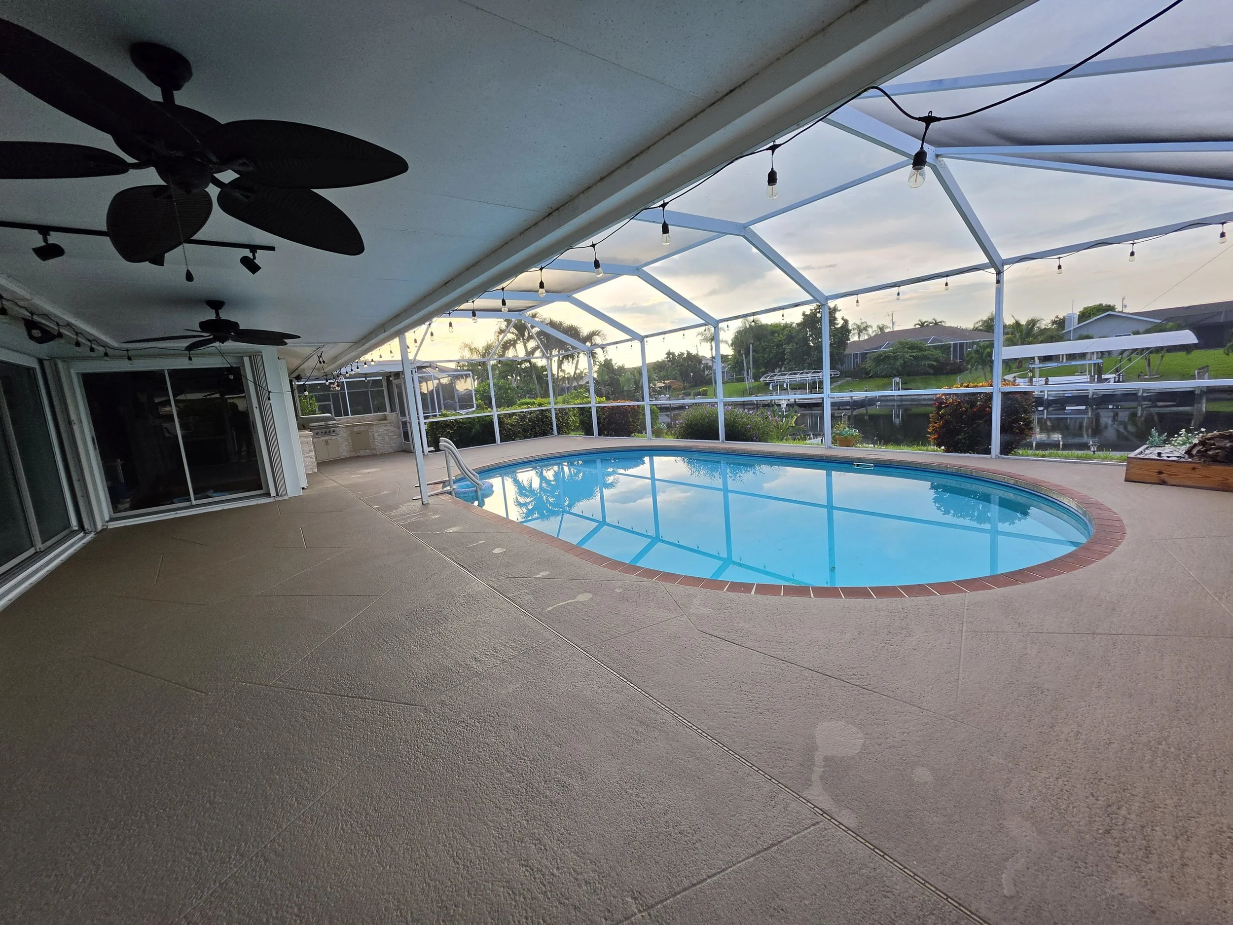 A backyard covered patio with ceiling fans, string lights, an in-ground pool, and a view of neighboring homes and greenery.
