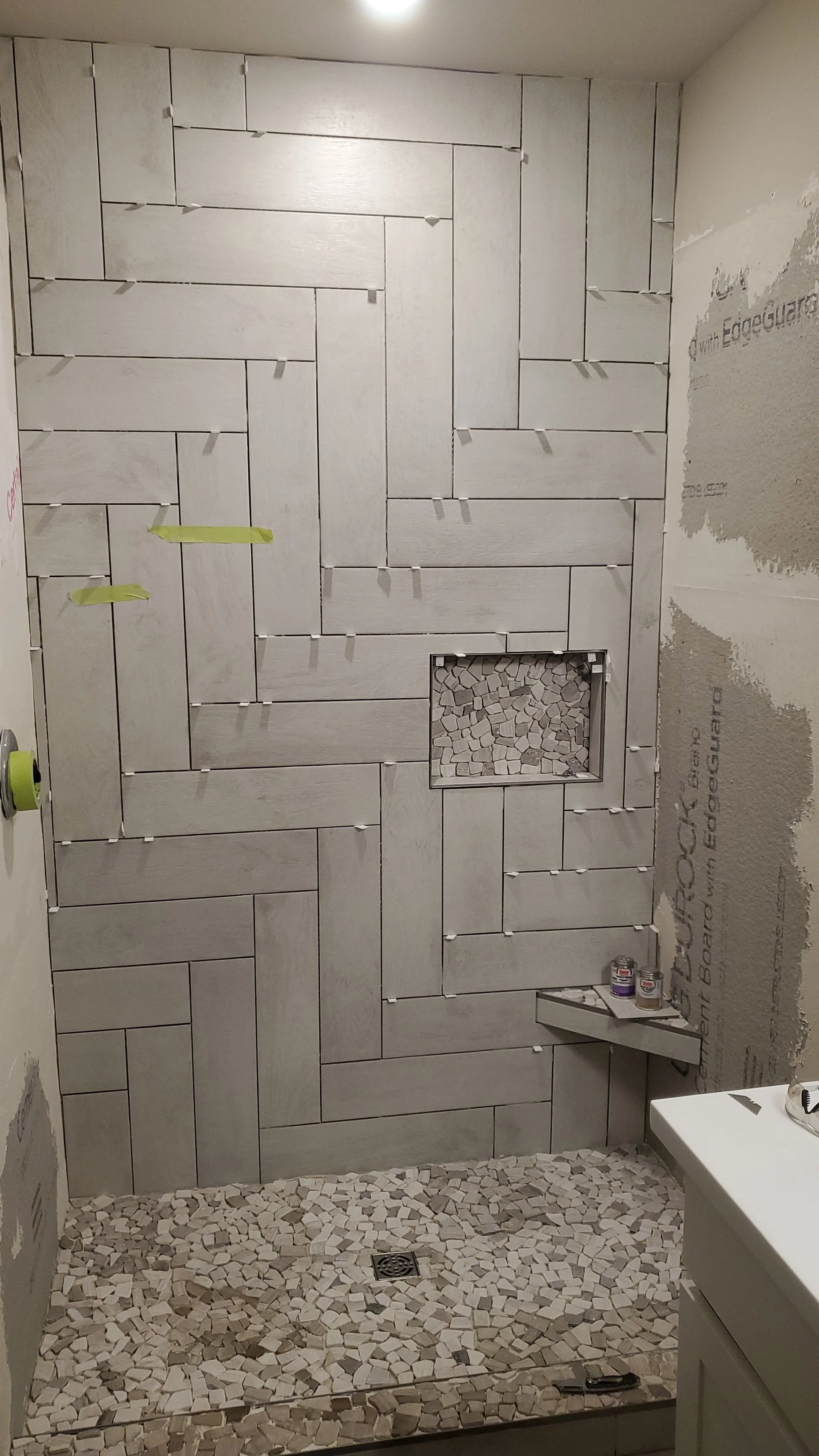 Bathroom under construction with tiled wall, mosaic floor, and a niche filled with small tiles, with construction supplies on a shelf.