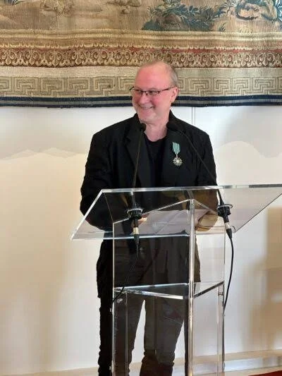 A smiling man with glasses and a black suit standing behind a clear podium, speaking in a room with a decorative tapestry on the wall.