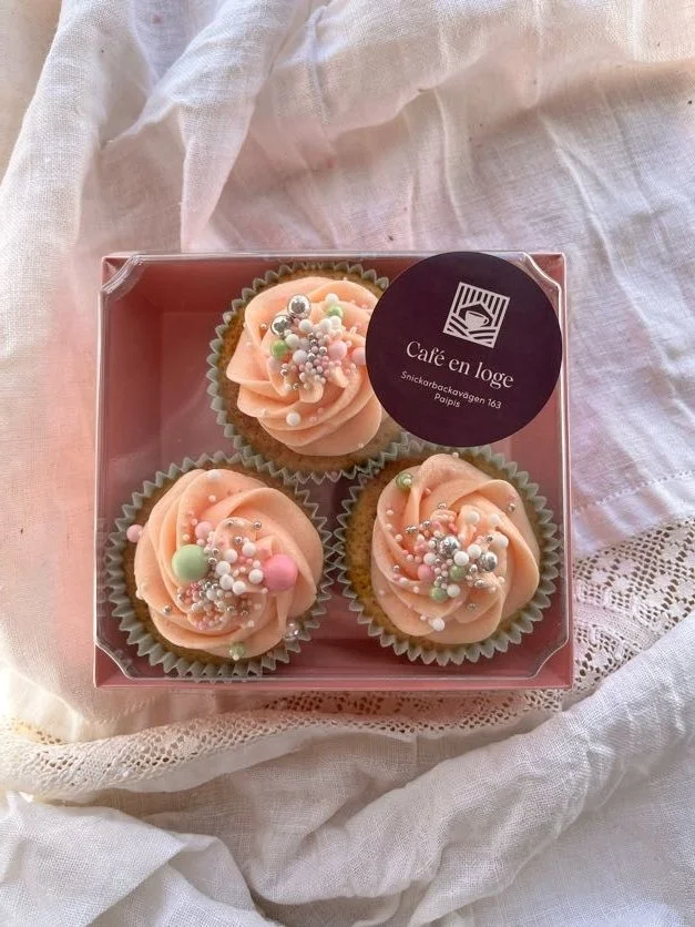 Box of three cupcakes with pink frosting and colorful sprinkles, in a pink box on a white fabric background.