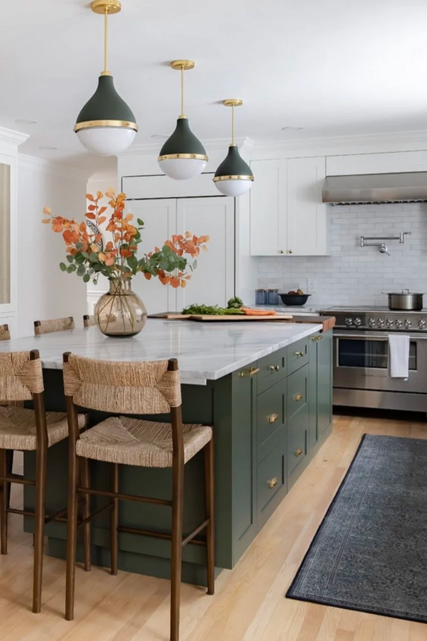 Modern kitchen with white cabinets, a marble island, three green pendant lights, a vase with orange leaves on the island, a stainless steel stove, and a gray area rug.
