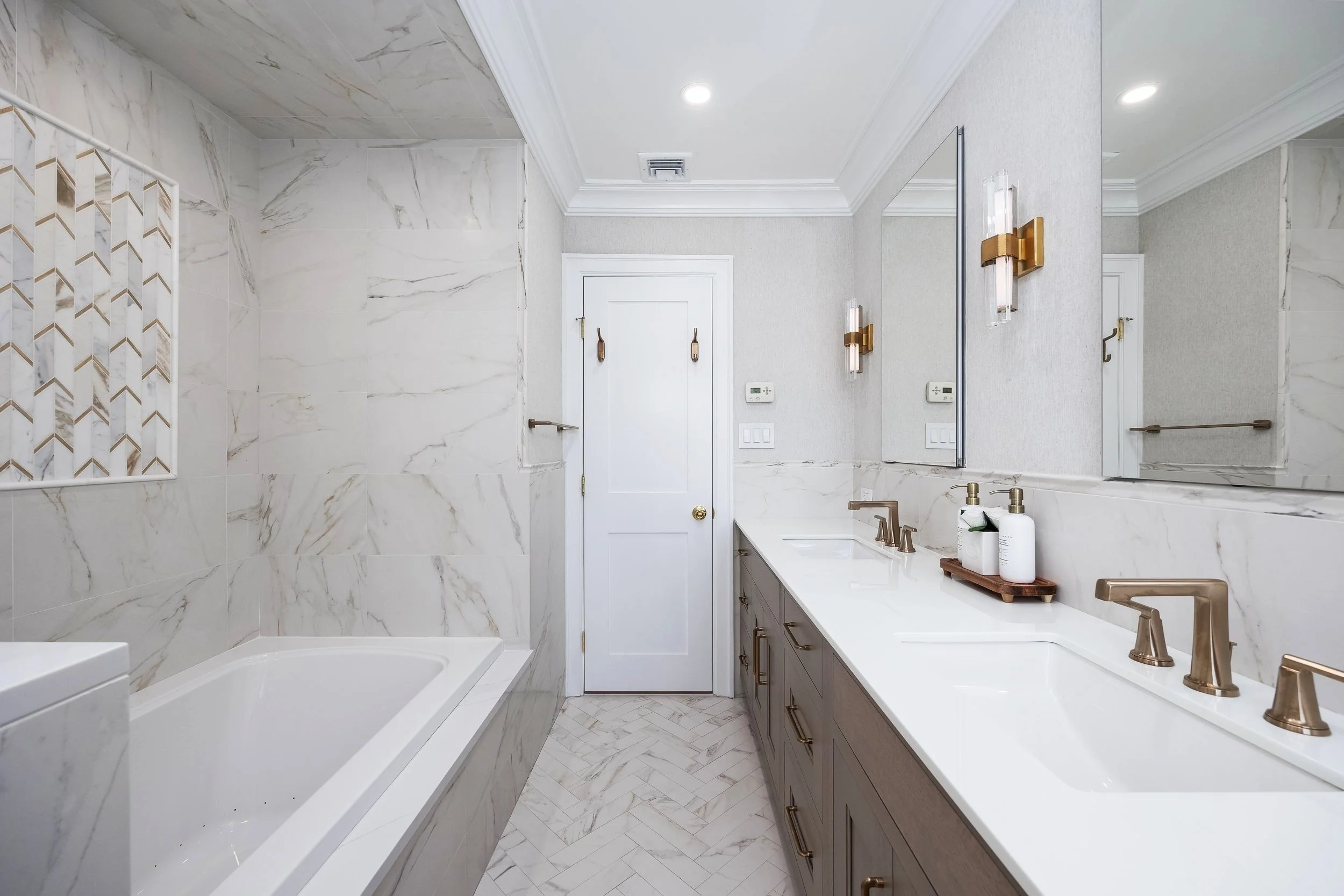 Modern bathroom with marble walls and floors, large mirror, double sink vanity with brass fixtures, wall-mounted light fixtures, and a white door.