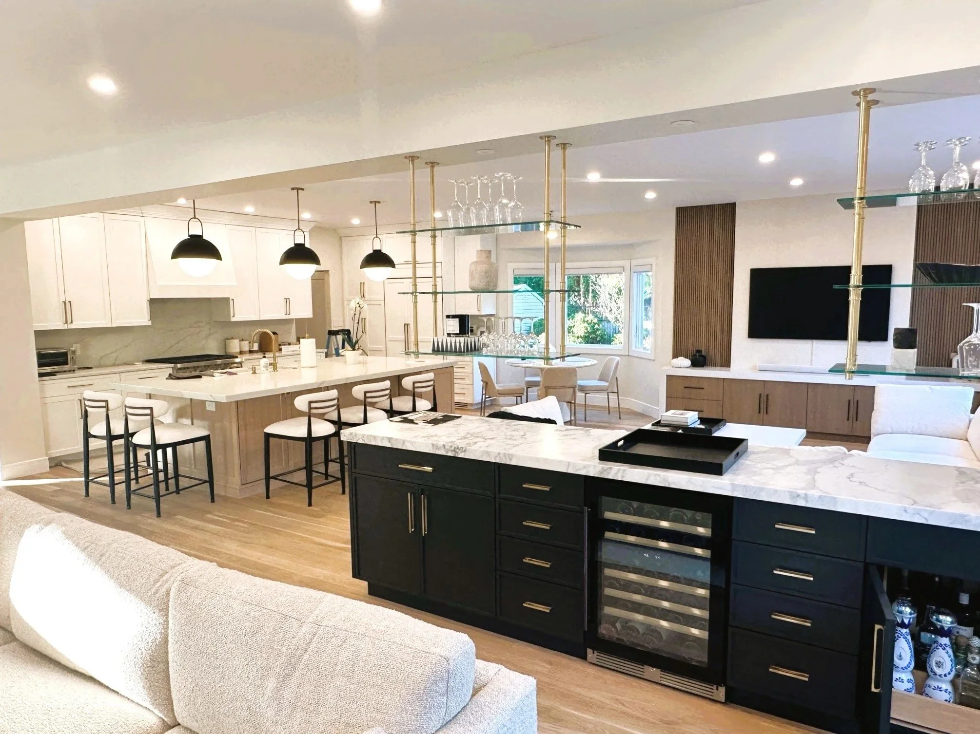 Modern open-concept kitchen with white cabinets, marble countertops, and black pendant lights, adjacent to a dining area with round table and chairs, and a living space with a beige sofa, featuring large windows and a mounted flat-screen TV.