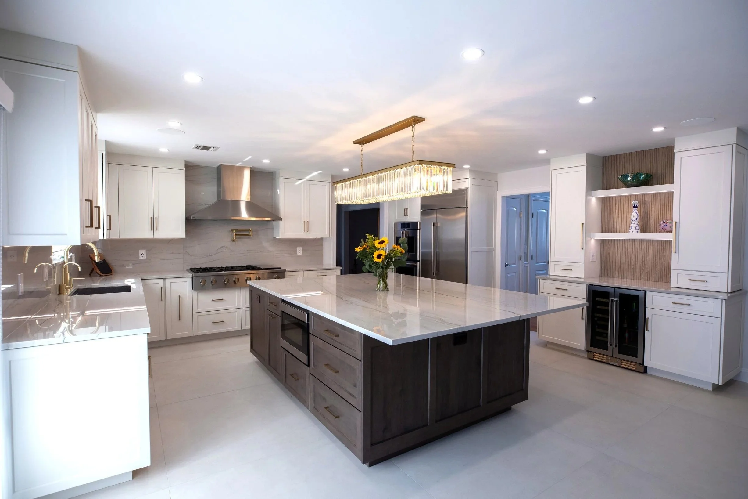 Modern kitchen with white cabinets, a large island with a dark base and marble countertop, stainless steel appliances, a central chandelier, and a sink with a gold faucet.